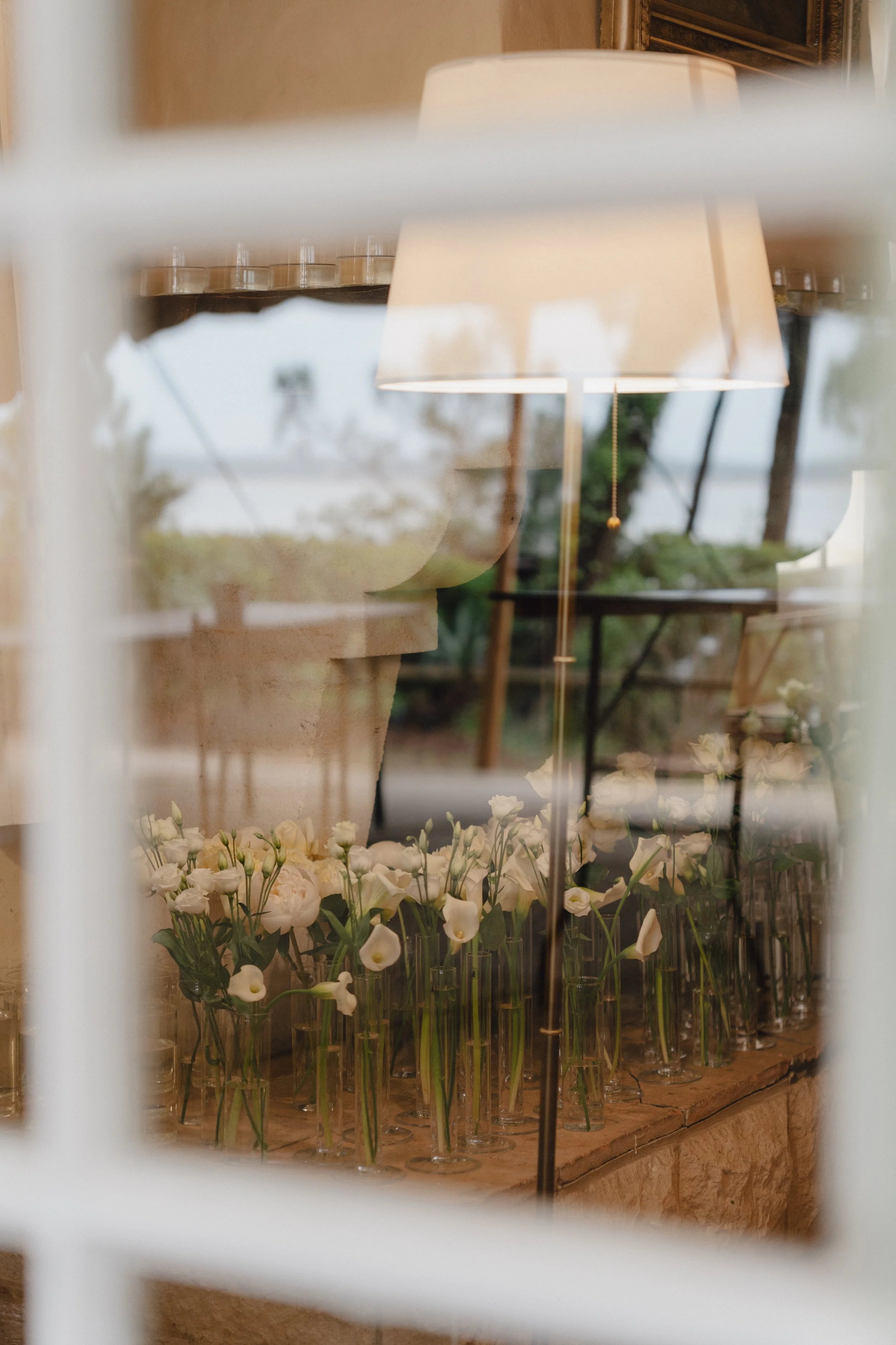 Vue à travers une fenêtre floue montrant des fleurs blanches dans des vases en verre, une lampe de table et un miroir en arrière-plan, dans une ambiance de décoration intérieure élégante.