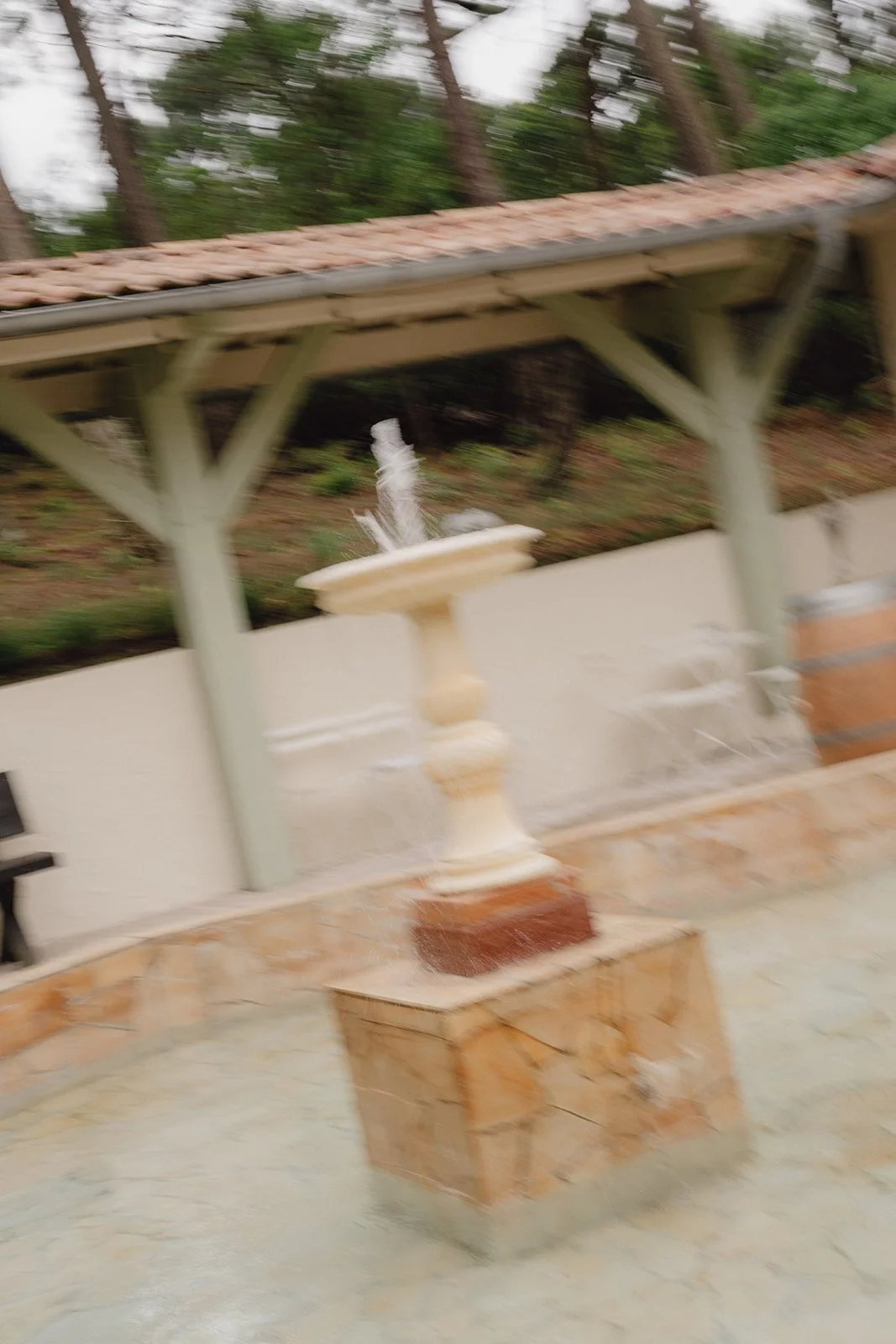 Une fontaine en pierre blanche avec de l'eau qui coule, située dans un espace extérieur avec un pavage en marbre beige et une clôture en bois, sous un toit en tuiles brunes, avec des arbres verts en arrière-plan. Photo floue, moderne.
