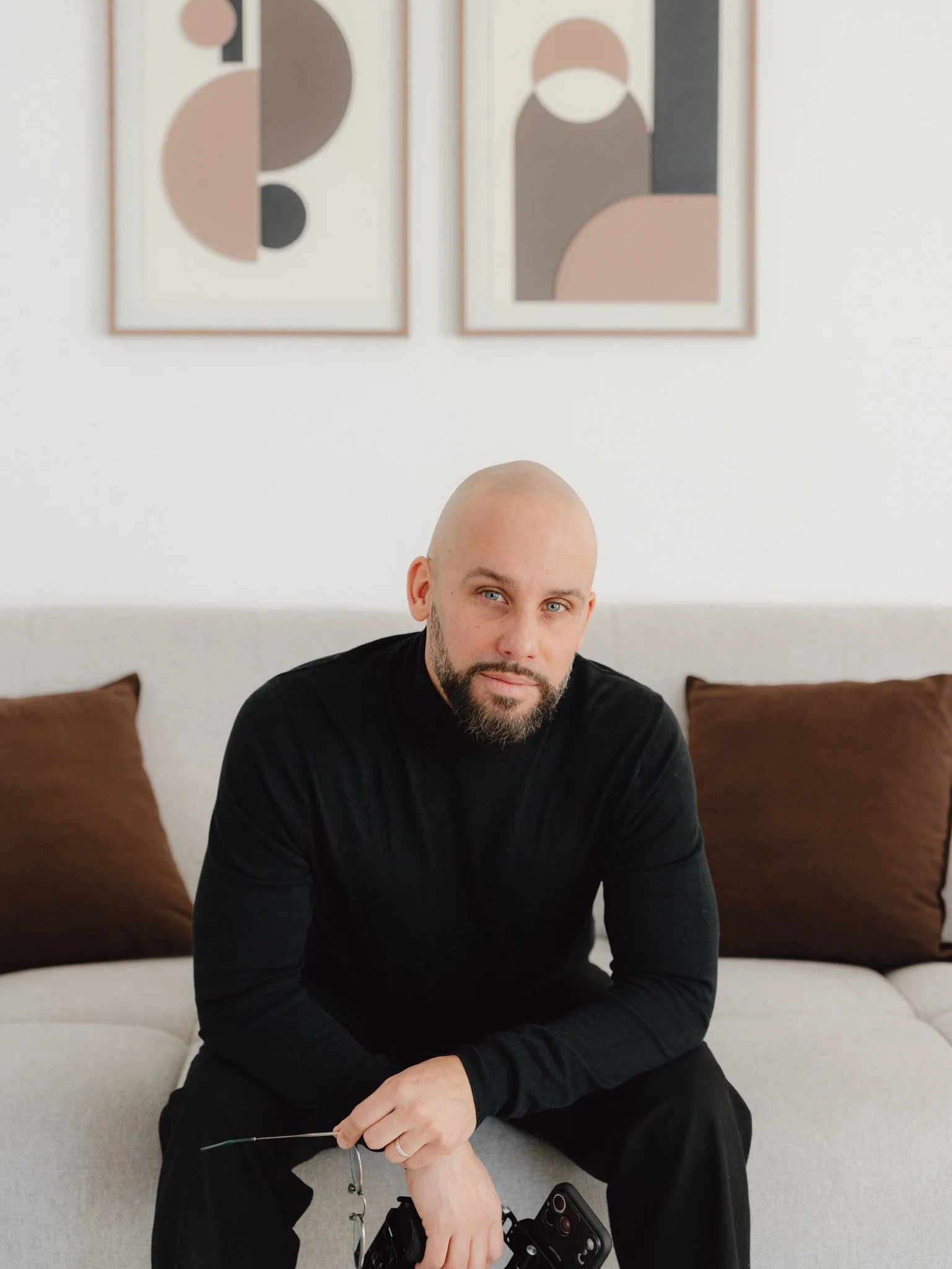 Portrait homme avec une tête rasée, portant un pull noir, assis sur un canapé beige avec deux coussins marron, dans un intérieur moderne avec deux tableaux abstraits au mur en arrière-plan.