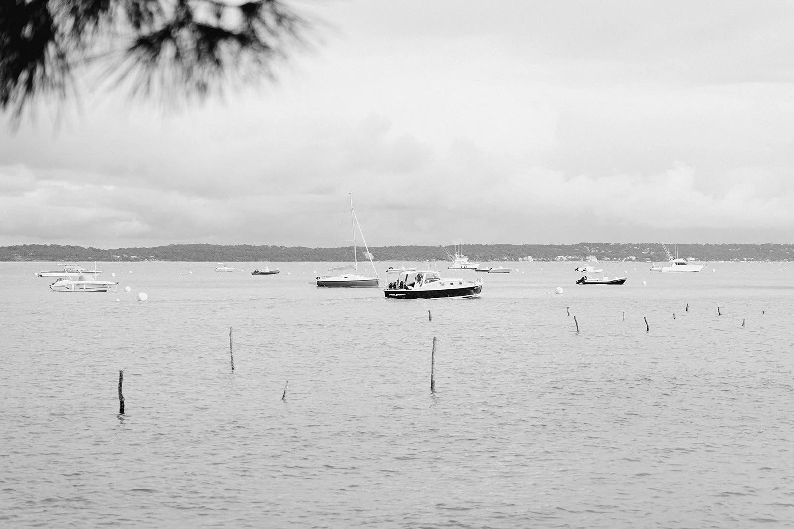 Vue en noir et blanc de bateaux à l'ancre sur une mer calme, avec un ciel nuageux et une côte au loin.