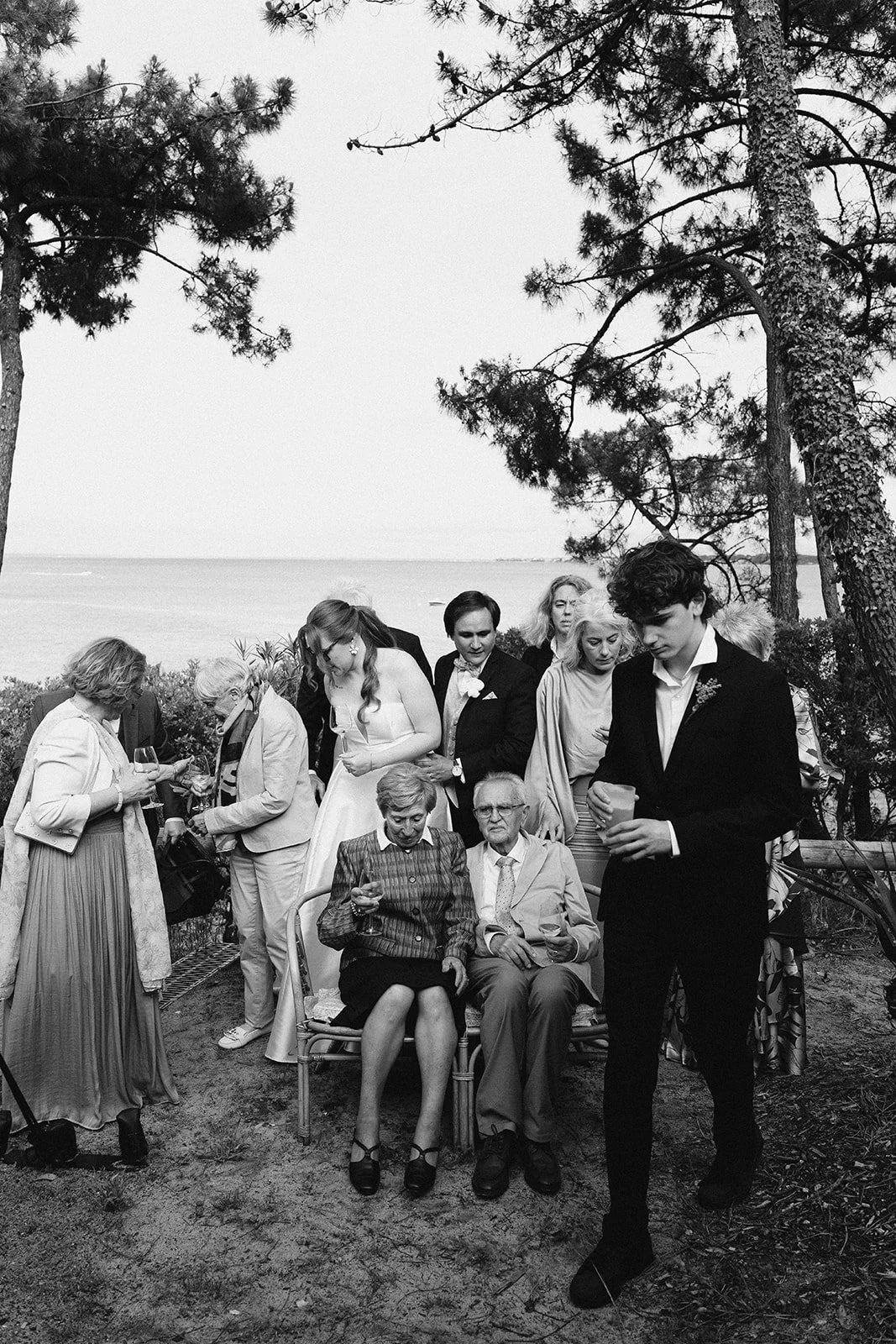 Groupe de personnes lors d'une célébration en plein air près de la mer, en noir et blanc, avec des arbres en arrière-plan.