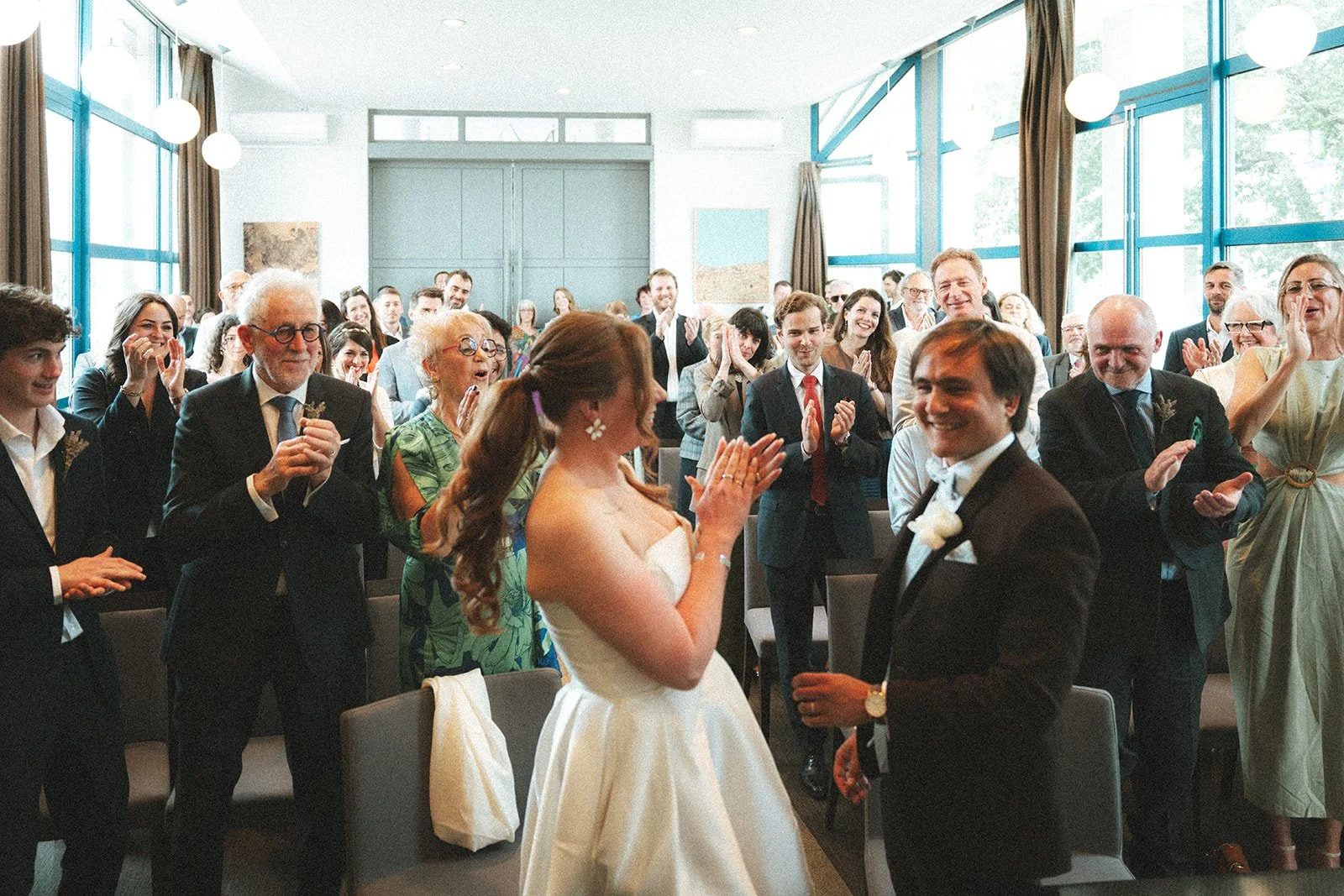 Un couple de mariés dans une salle de mariage, entourés de plusieurs invités qui applaudissent et sourient.