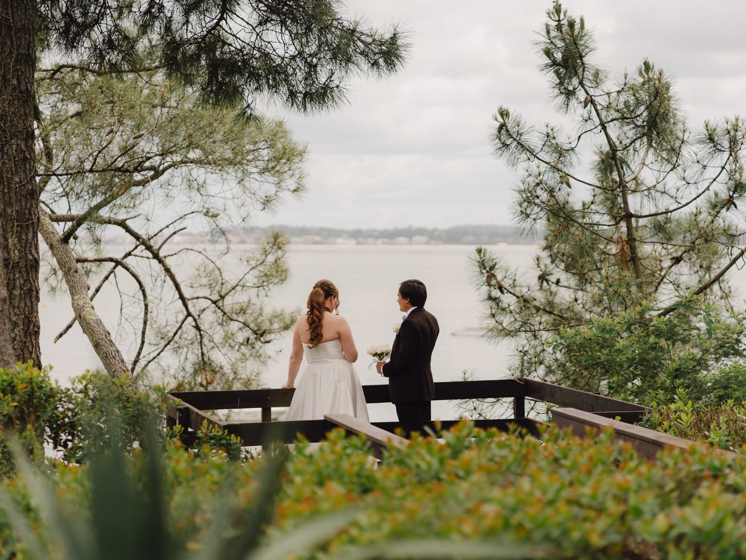 Un couple en costume de mariage échange des vœux devant un devant un paysage naturel avec des arbres et un lac, sous un ciel nuageux.