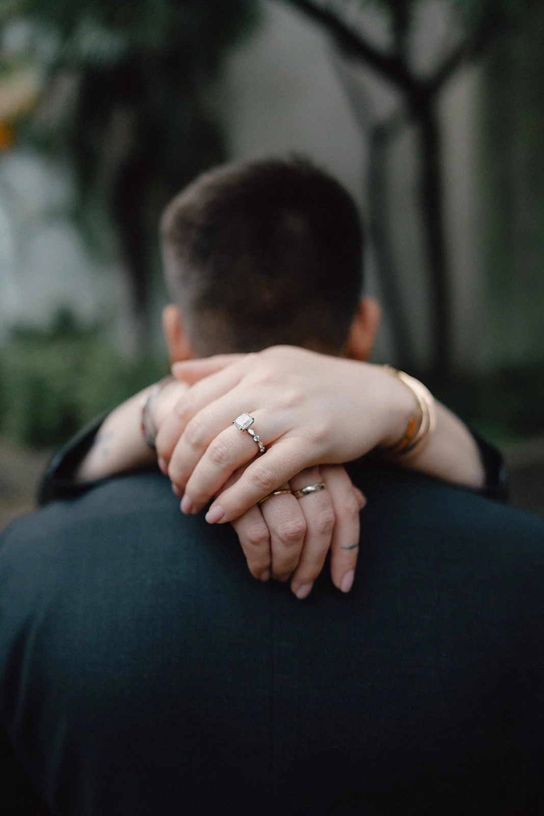 Deux personnes avec des ongles longs et décorés, s'embrassent de derrière, avec un fond flou de végétation.