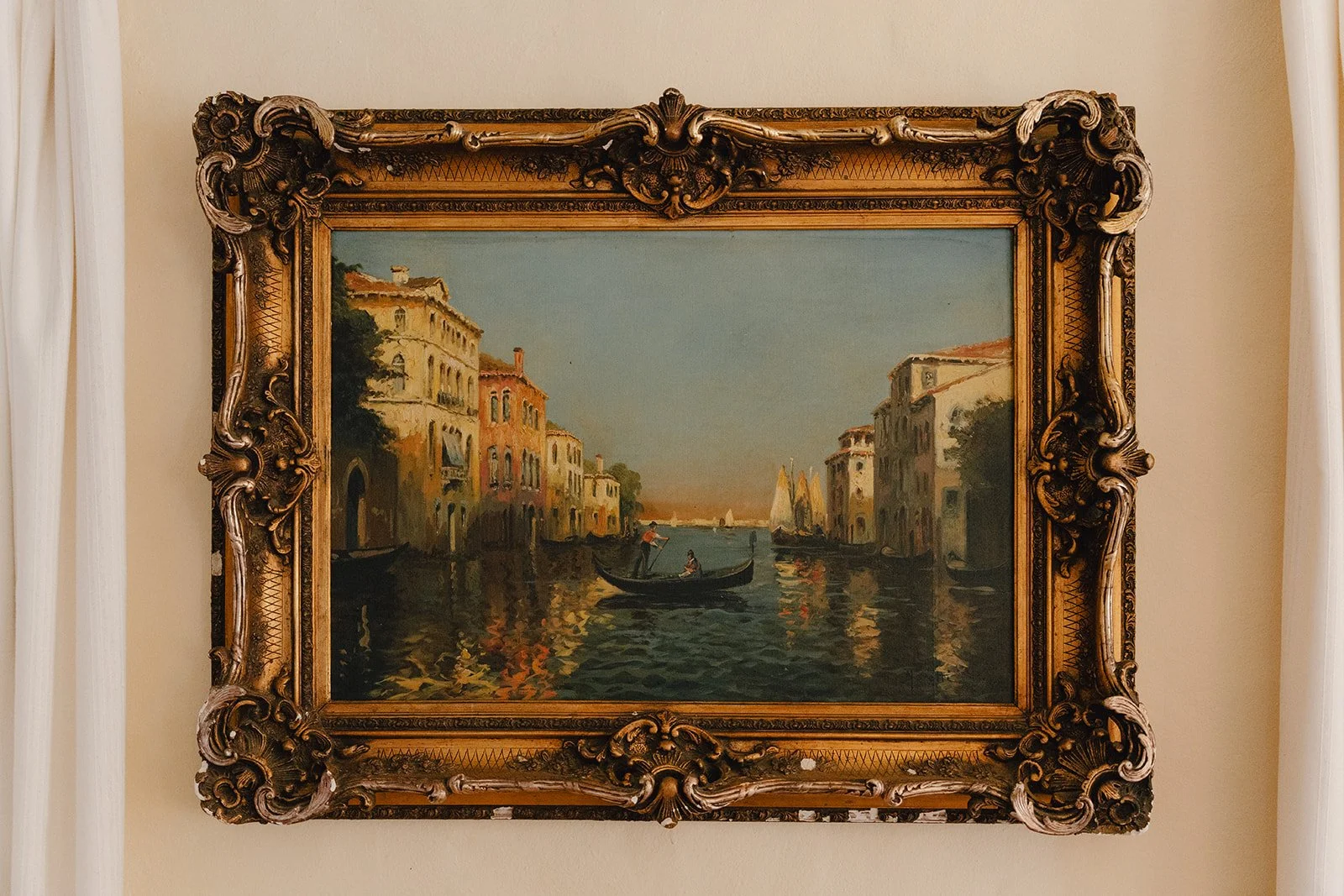 Peinture représentant une scène de canal à Venise avec des bâtiments anciens, des bateaux et des voiliers, encadrée d'un cadre en bois orné de sculptures délicates.