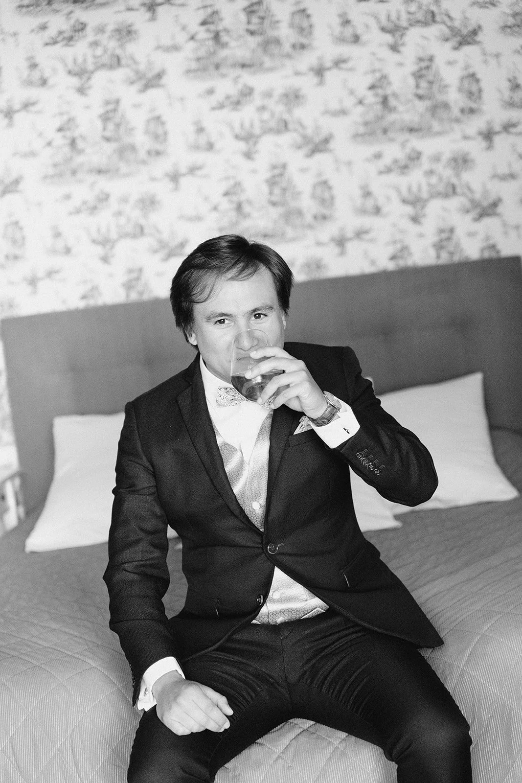Un homme habillé en costume noir, assis sur un lit, en train de boire un verre, séance portrait marié.