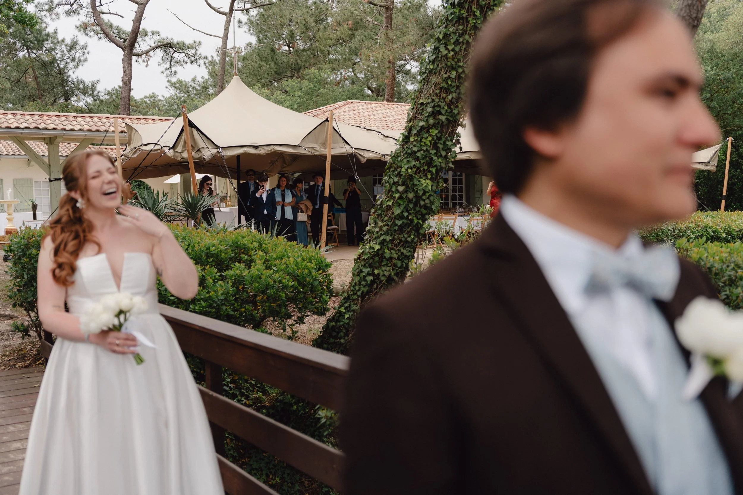 Le first look d'un couple de mariés avec des invités en arrière-plan, arborant une tente et des arbres autour. La mariée est tout sourire, chez le marié, la tension monte.