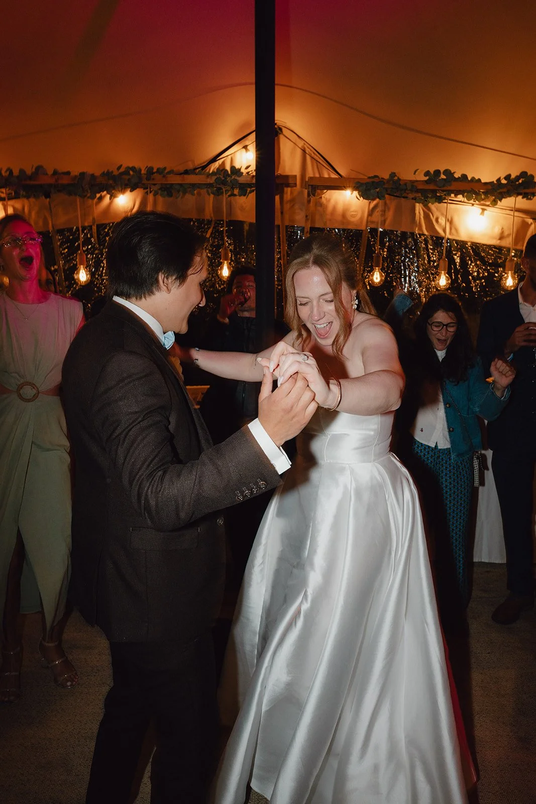 Un couple danse lors de leur mariage, entouré d'amis, sous un éclairage chaleureux dans une tente décorée.