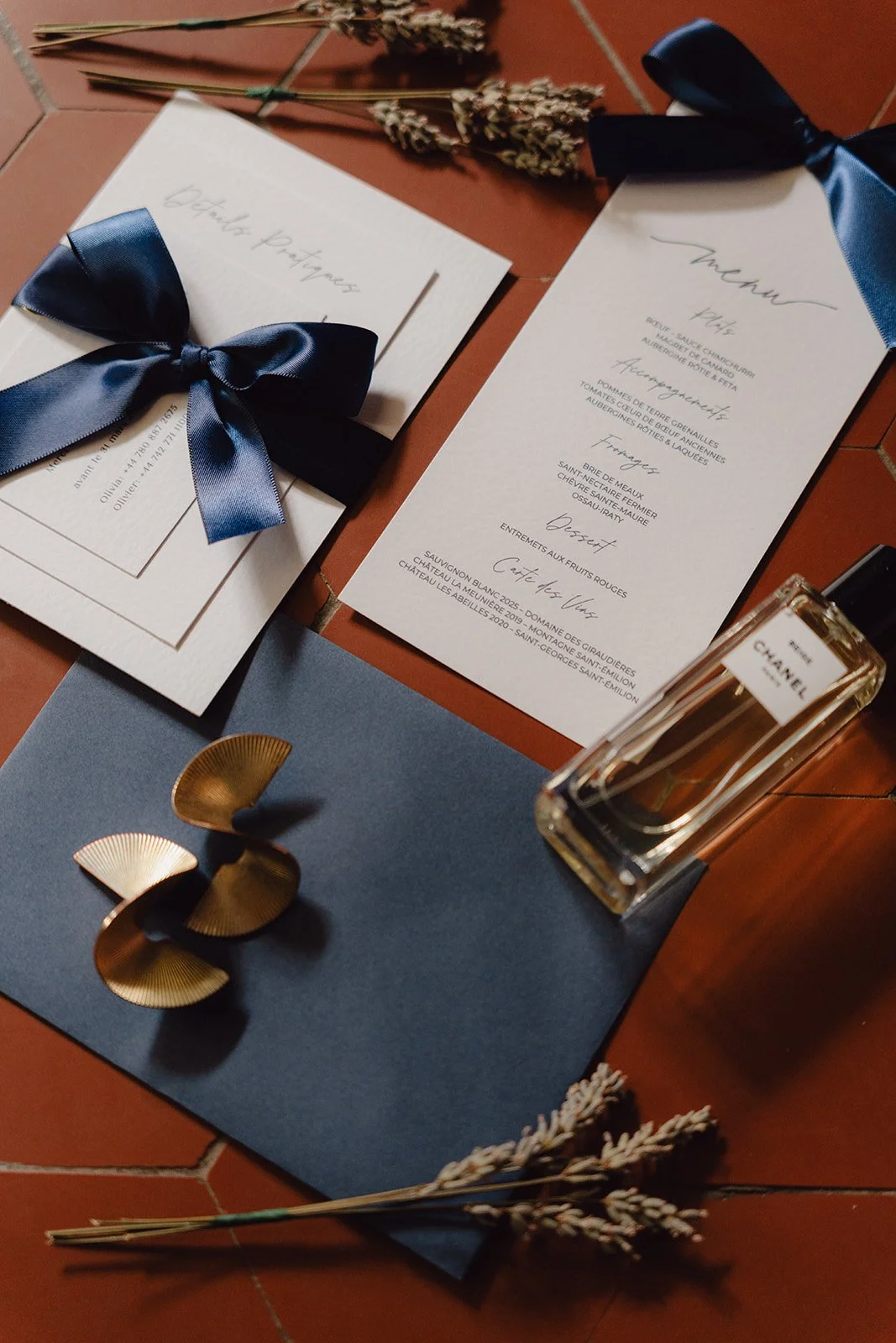 Flatlay du Menu de dîner, une bouteille de parfum Chanel, un bouquet de lavande, un tapis de table bleue avec des pièces décoratives en or, un livret, un carton avec un ruban en satin bleu, tous disposés sur une surface en terre cuite.