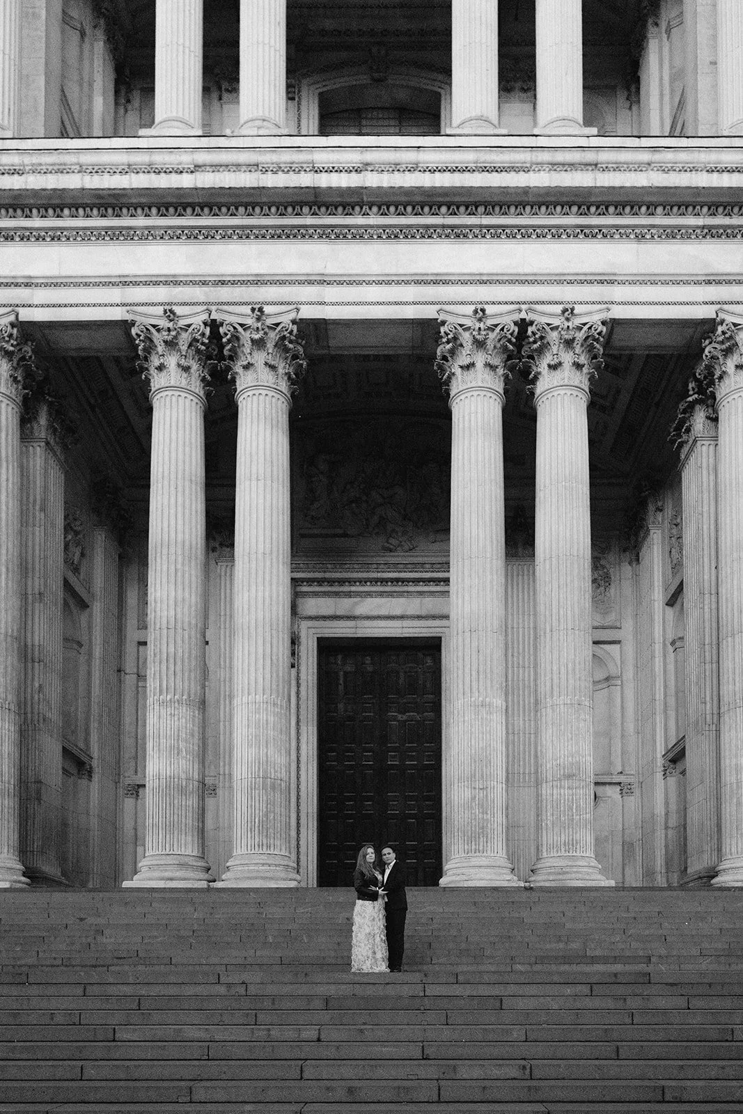 Deux personnes, un homme et une femme, se tiennent au centre des escaliers devant une grande façade classique ornée de colonnes ioniques.
