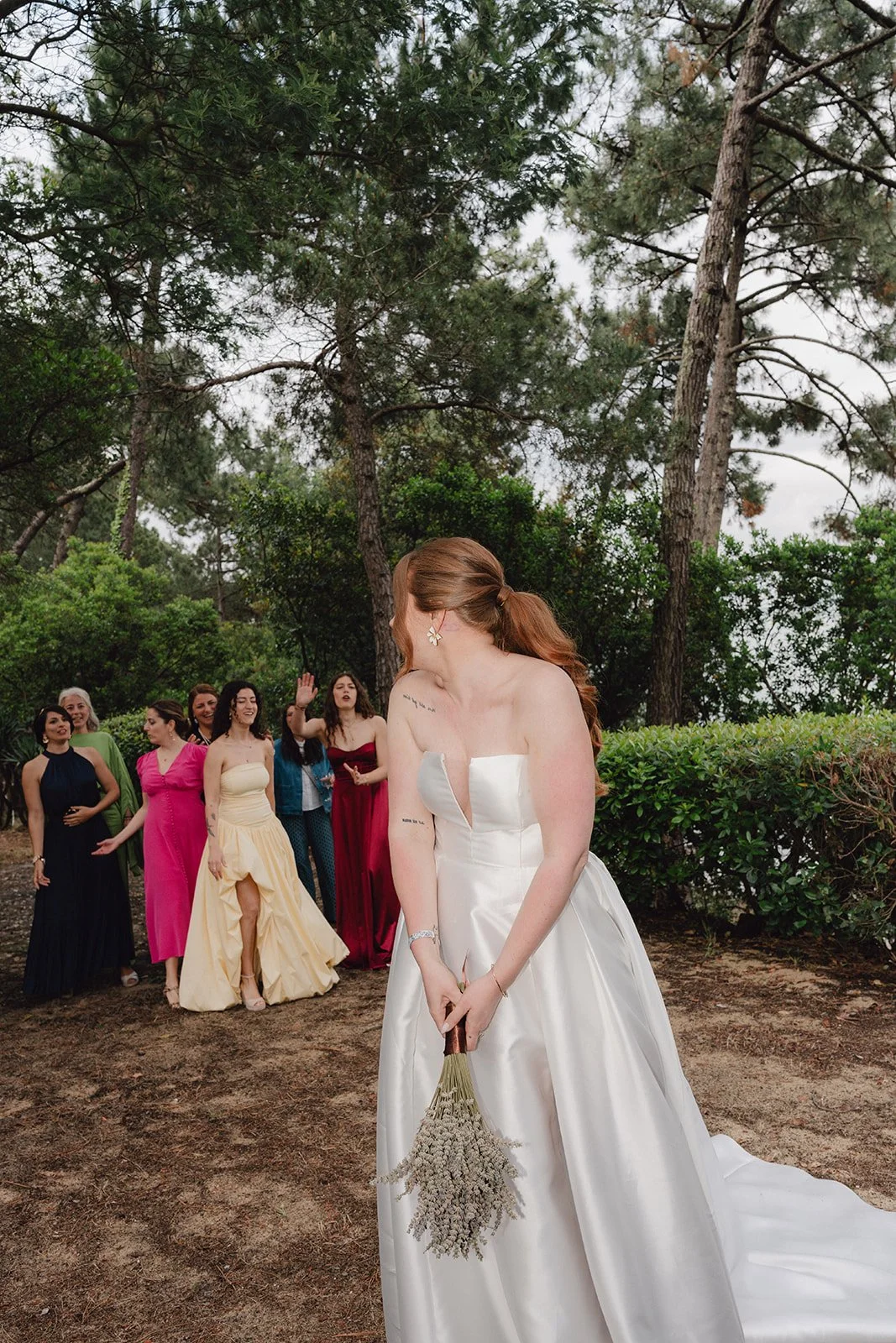 Une femme en robe de mariée blanche tient un bouquet de fleurs, tournant la tête pour éviter le regard d'un groupe de femmes en robes colorées, dans un cadre naturel avec des arbres.