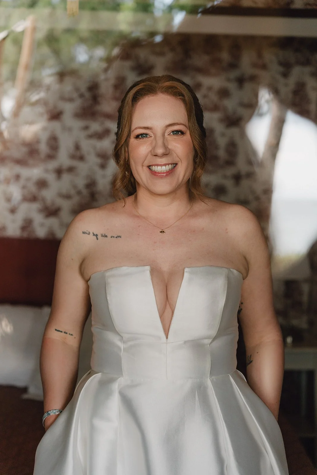 Une femme souriante portant une robe de mariage blanche, avec des tatouages visibles sur ses bras et ses épaules, dans une pièce avec un mur de pierre en arrière-plan.