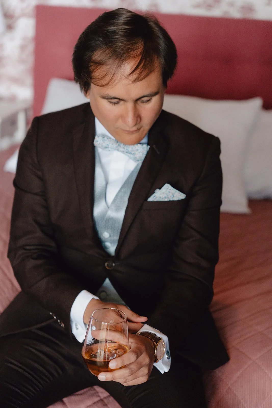 Un homme habillé en smokings noir et gris assis sur un lit, tenant un verre de vin rosé, regardant vers le bas, avec un fond de mur rose et un coussin blanc.