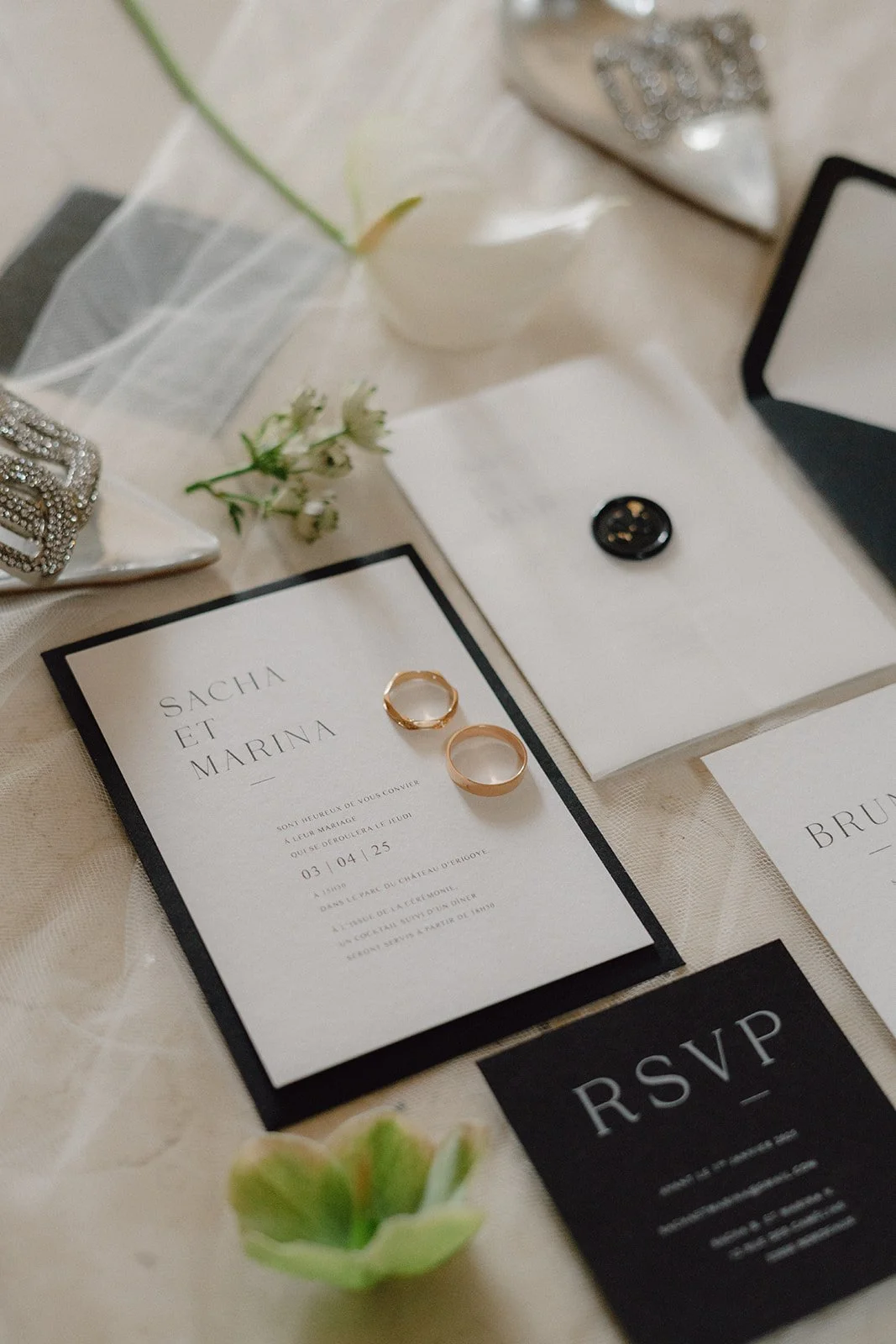 Flatlay papeterie noire et blanche de mariage avec deux alliances, fleurs blanches et éléments décoratifs par terre.