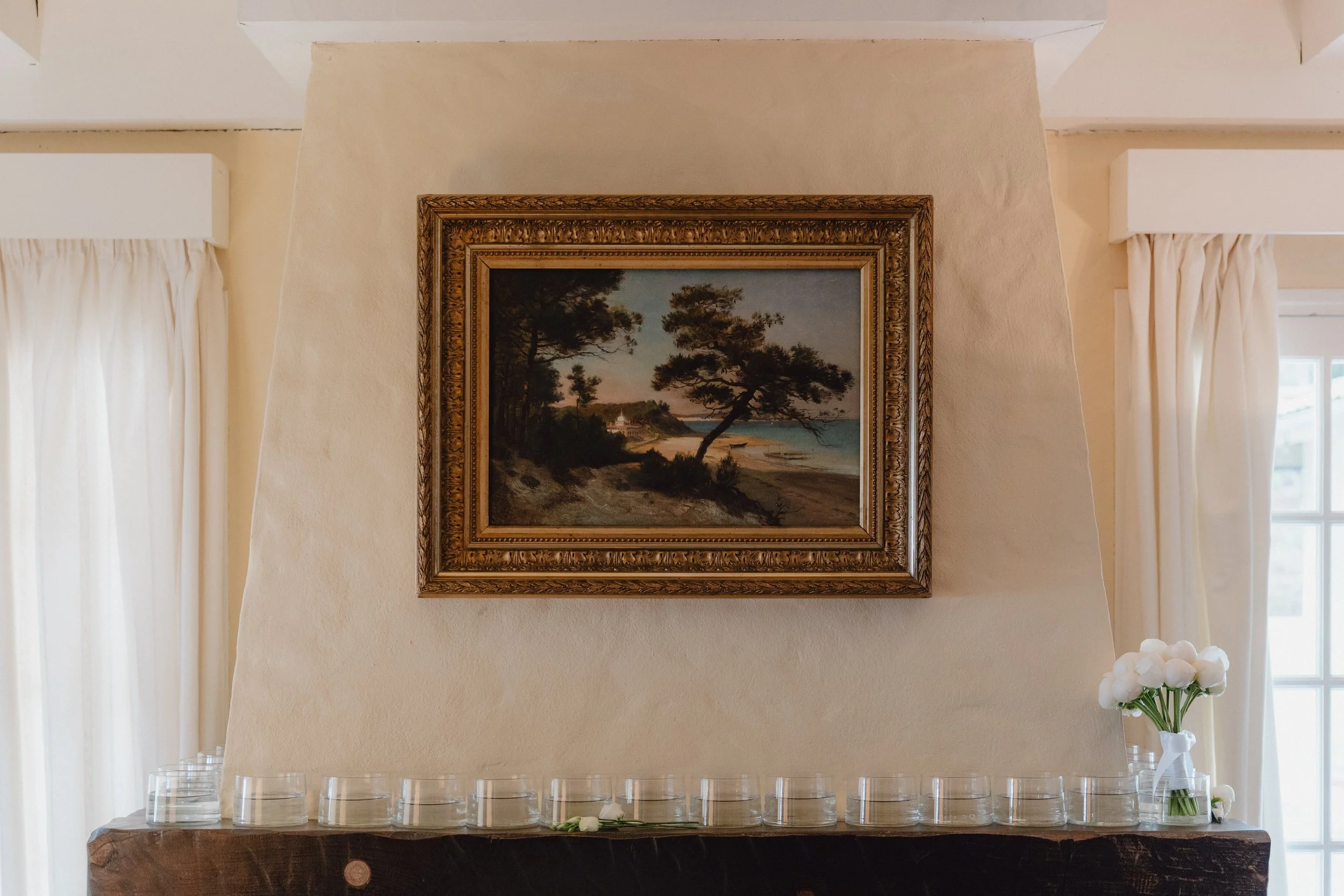 Une peinture représentant un paysage côtier avec un arbre tortueux, la mer et un coucher de soleil, dans un cadre doré, suspendue sur un mur beige près de fenêtres avec rideaux blancs, et un bouquet de fleur de mariage dans un coin.