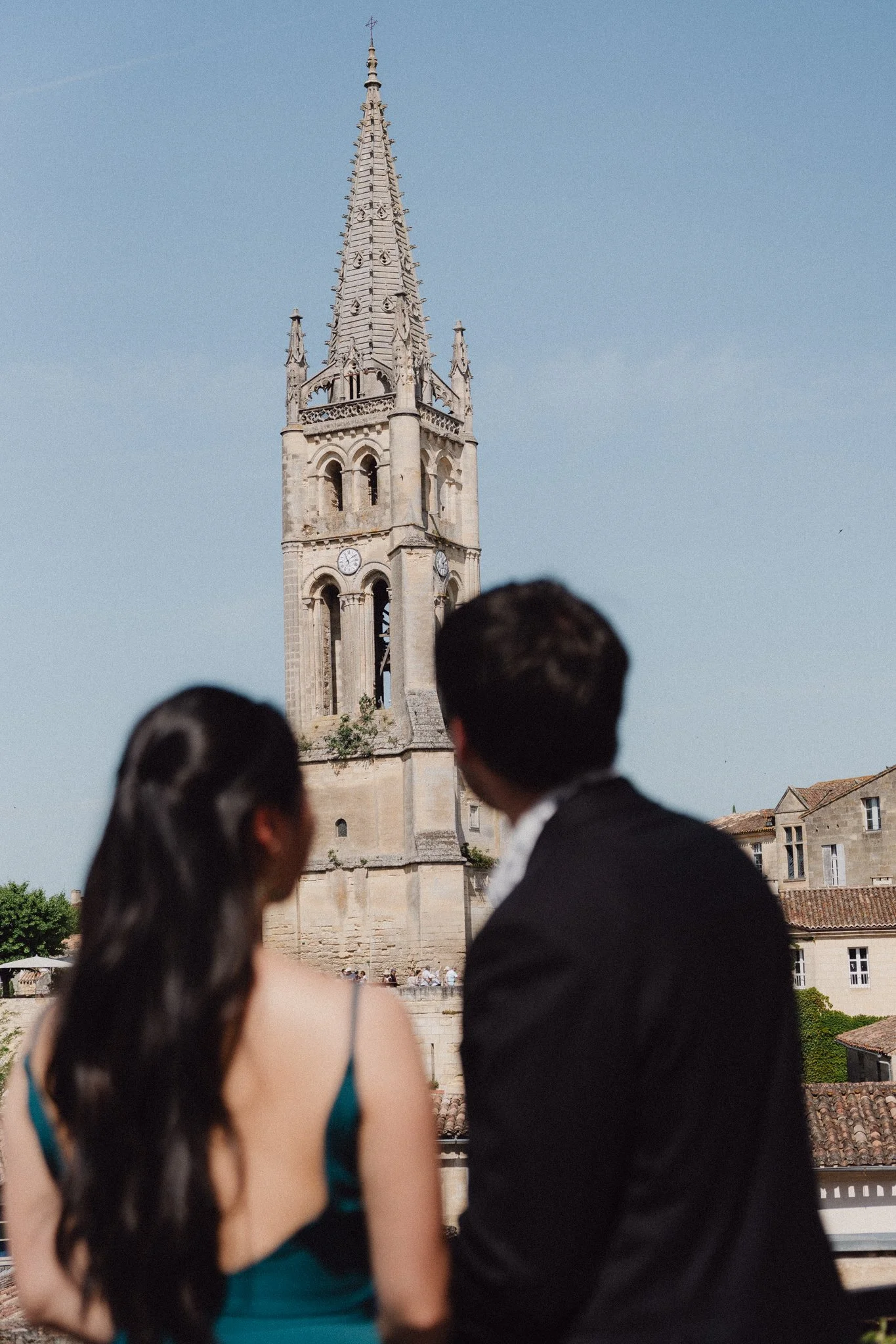 Une tour d'horloge ancienne sous un ciel bleu, avec trois personnes floues en premier plan, une femme avec de longs cheveux noirs, un homme en costume noir et une autre personne dont seule la tête est visible.