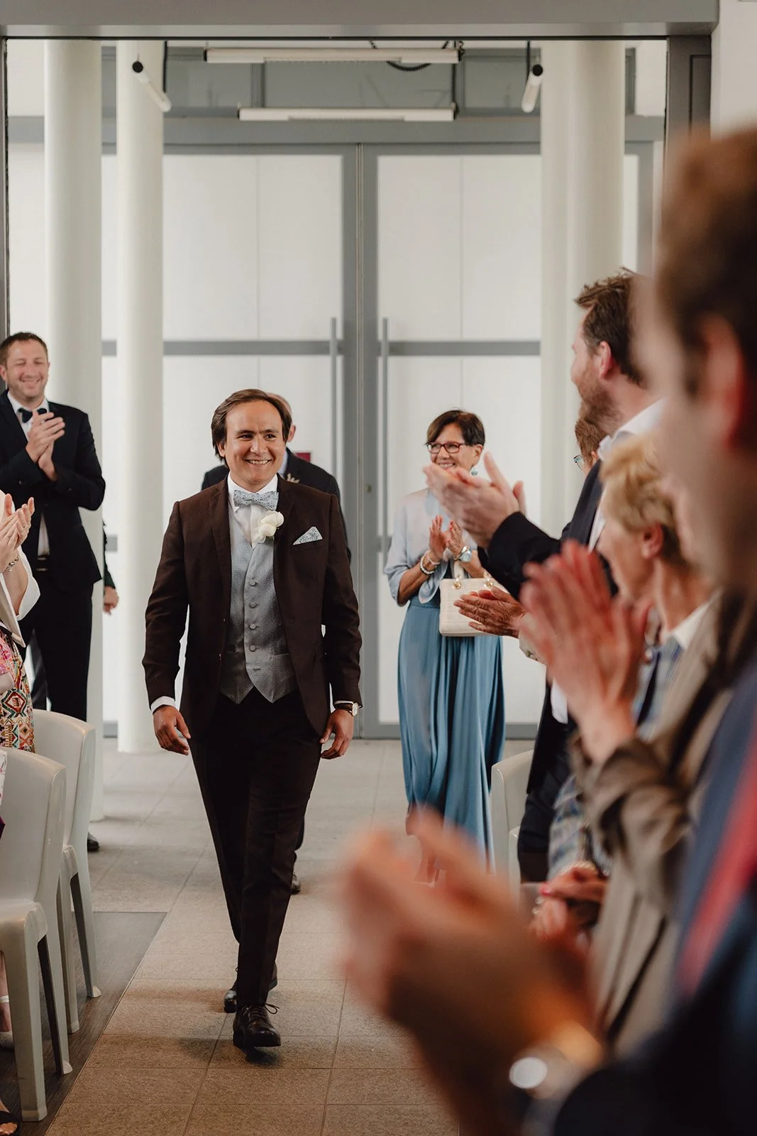 Un homme en costume de mariage marche dans une salle de cérémonie, entouré de personnes applaudissant, dans un décor moderne et clair.