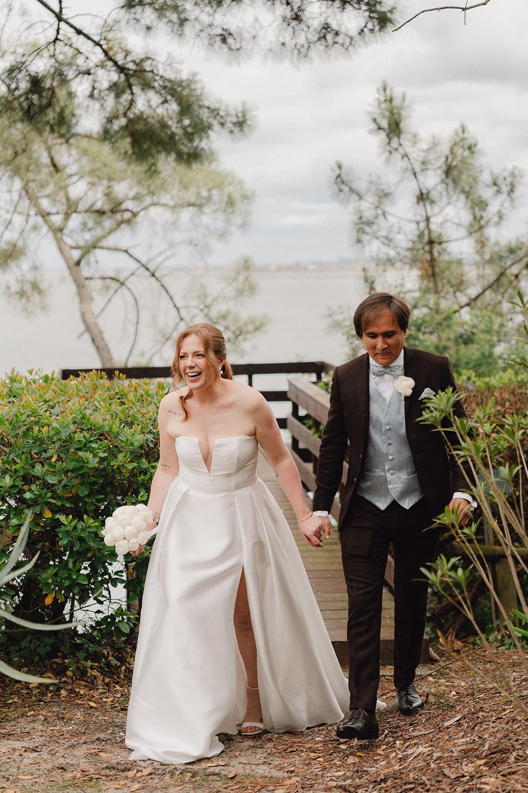 Un couple de mariés marche main dans la main dans un cadre naturel, elle en robe de mariage blanche, lui en costume sombre, entourés de verdure et près d'un lac.