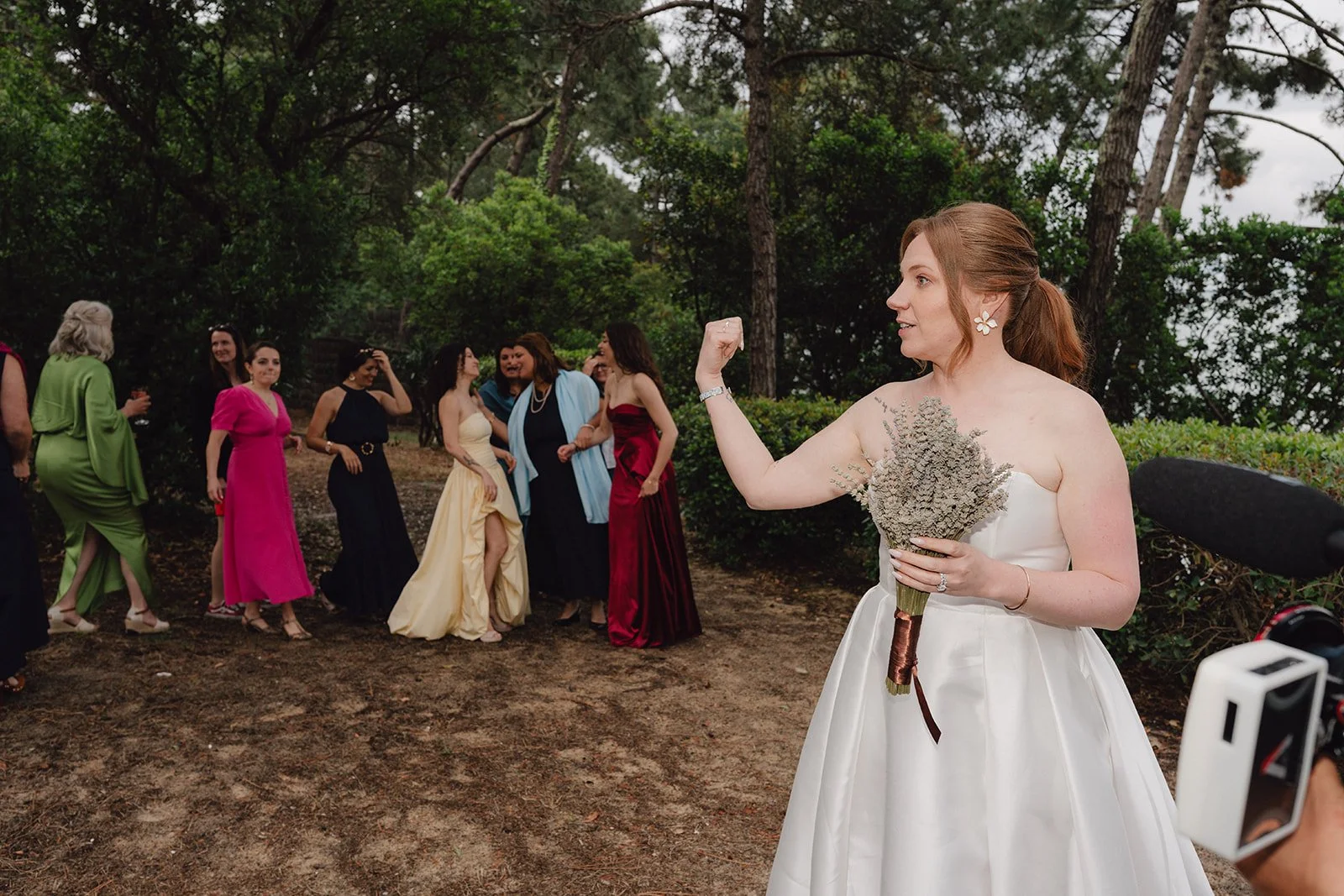 Une mariée en robe blanche, tenant un bouquet de fleurs, fait un geste de bras musclé vers un groupe de femmes en robes colorées lors d'une fête en extérieur, entourée d'arbres.