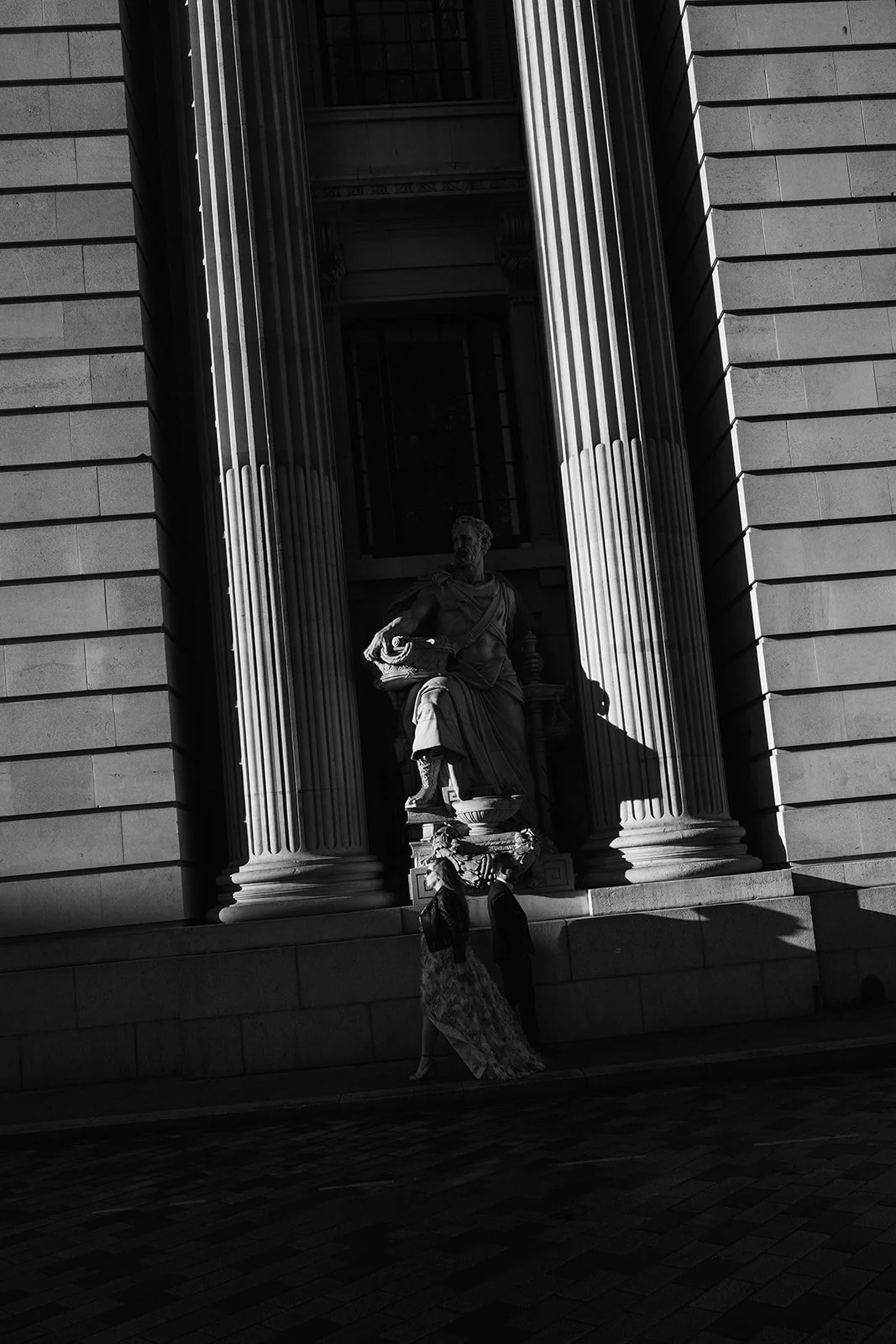 Une scène urbaine en noir et blanc montrant un bâtiment avec des colonnes et une statue d'une figure historique ou mythologique, avec deux personnes passant devant.