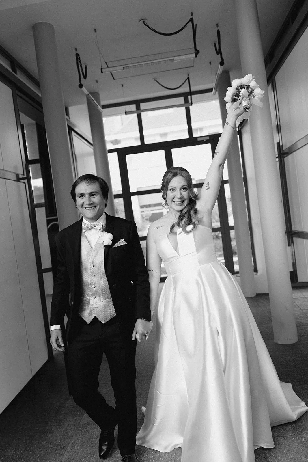Un couple de mariés pose à l'intérieur d'un bâtiment moderne, la mariée tient un bouquet en l'air, la robe blanche et le marié porte un costume noir avec une cravate en velours, sourire et ambiance joyeuse.