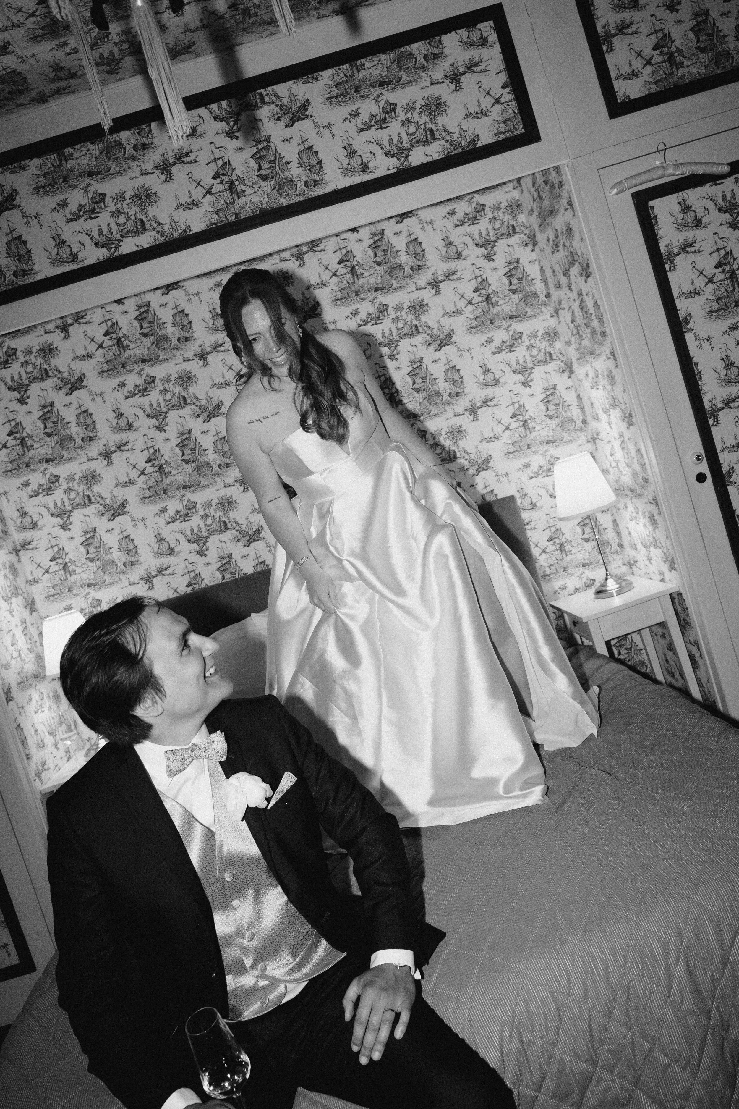 Séance photo couple, moment détente dans la chambre nuptiale, un verre de vin dans la main du marié, la mariée ravie de cette bulle et saute sur le lit.