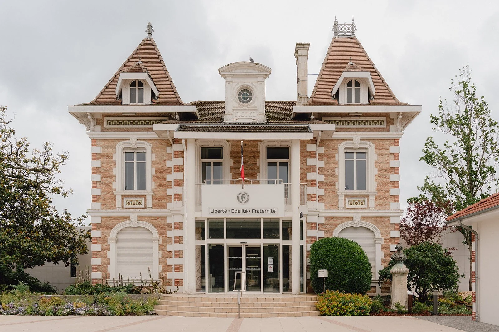 Bâtiment en briques rouges avec détails blancs, deux tours avec toits en pente, façade avec colonnes, drapeau français, inscription "Liberté, Égalité, Fraternité", jardins en avant.
