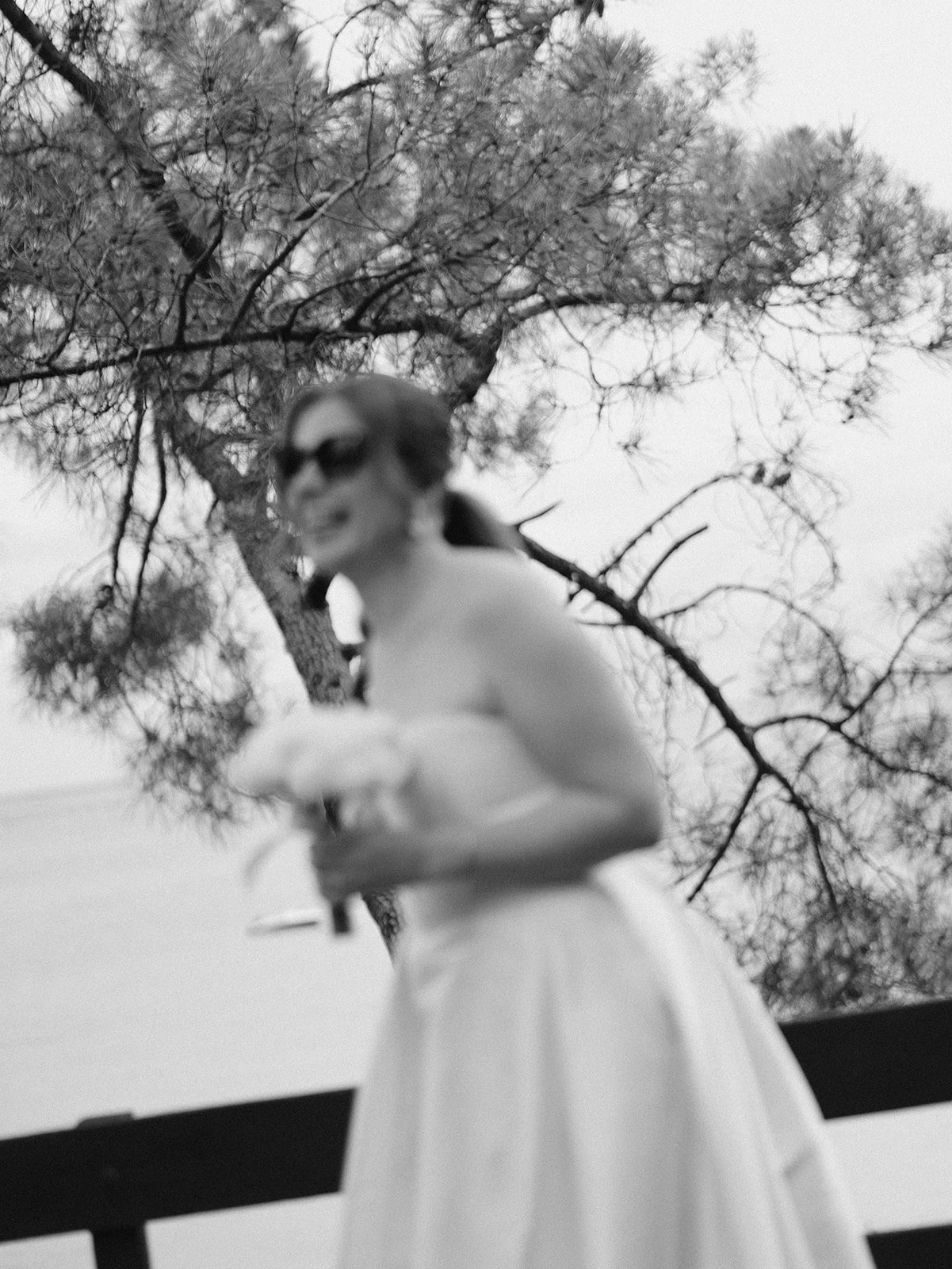 Une femme portant une robe blanche et des lunettes de soleil, tenant une coupe à la main, debout devant un arbre nu, en noir et blanc.