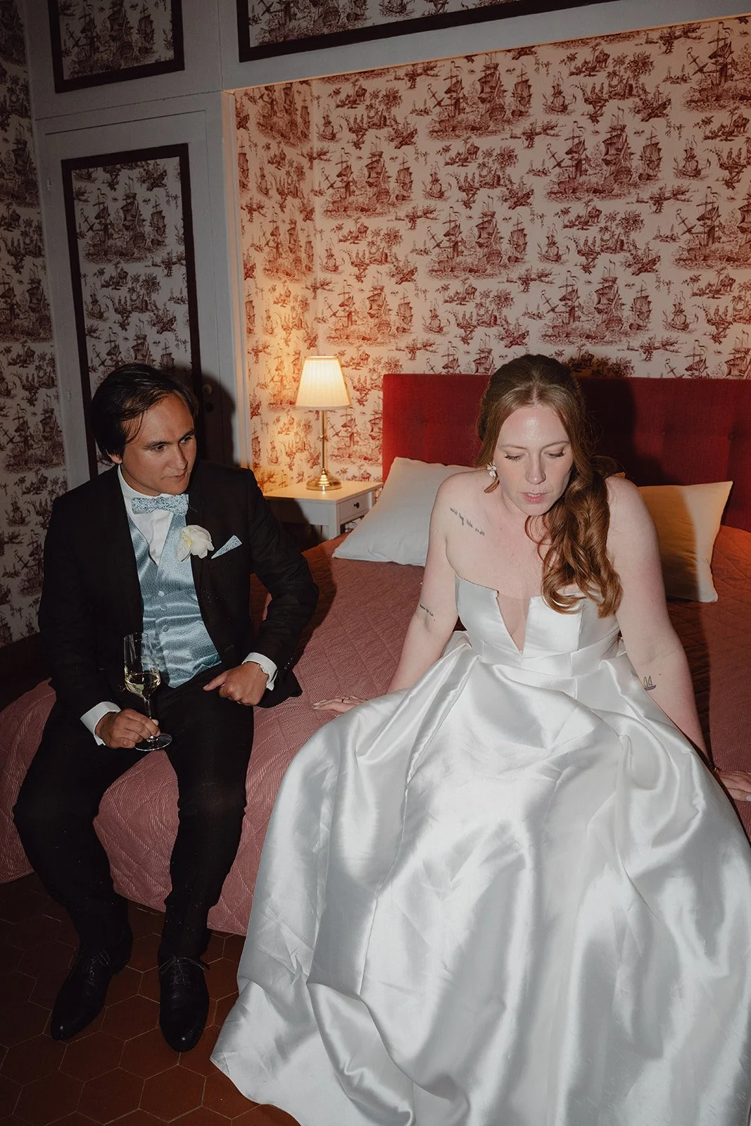 Séance couple au style éditorial. Un homme en smoking noir et une femme en robe de mariée blanche assis sur un lit, dans une chambre décorée avec du papier peint à motifs et une lampe de chevet.