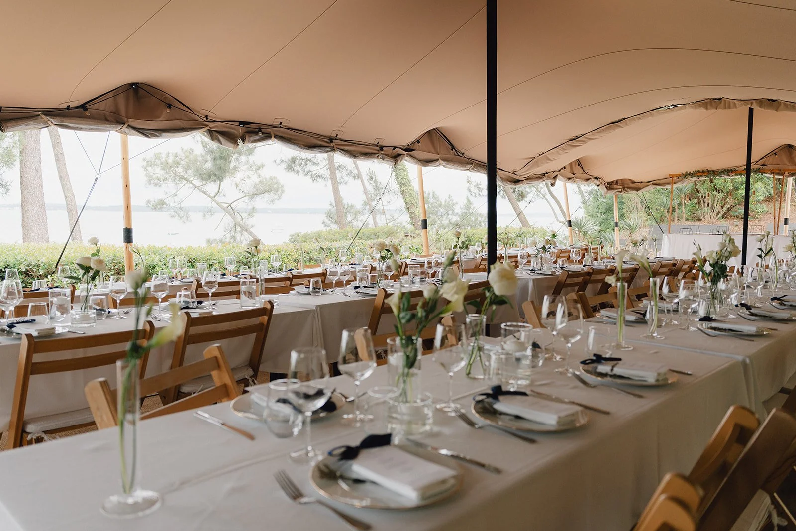 Une grande tente aménagée pour un événement ou un mariage, avec des tables longues et ornées de fleurs blanches, de vaisselle élégante et de verres, offrant une vue sur un espace extérieur avec des arbres et un plan d'eau en arrière-plan.