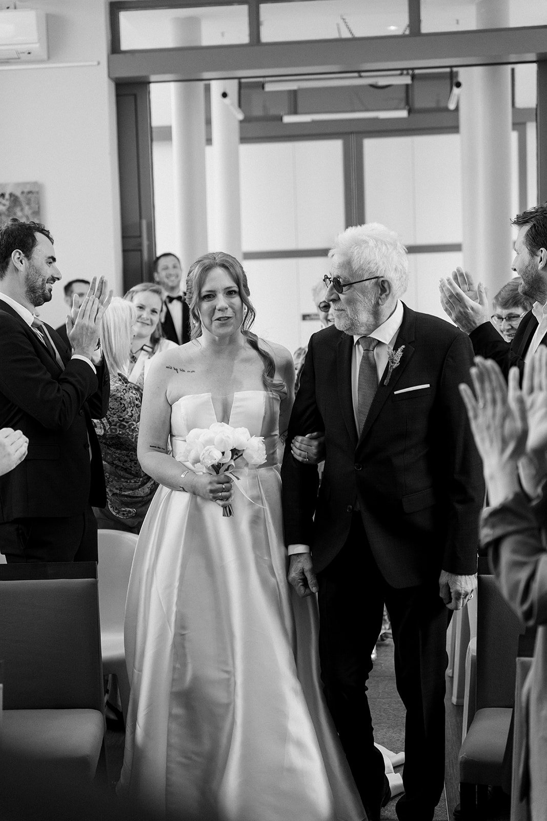 Une jeune femme en robe de mariage tenant un bouquet, marchant dans une salle lors d'une cérémonie, accompagnée d'un homme âgé en costume, entourée de personnes applaudissant et souriant.