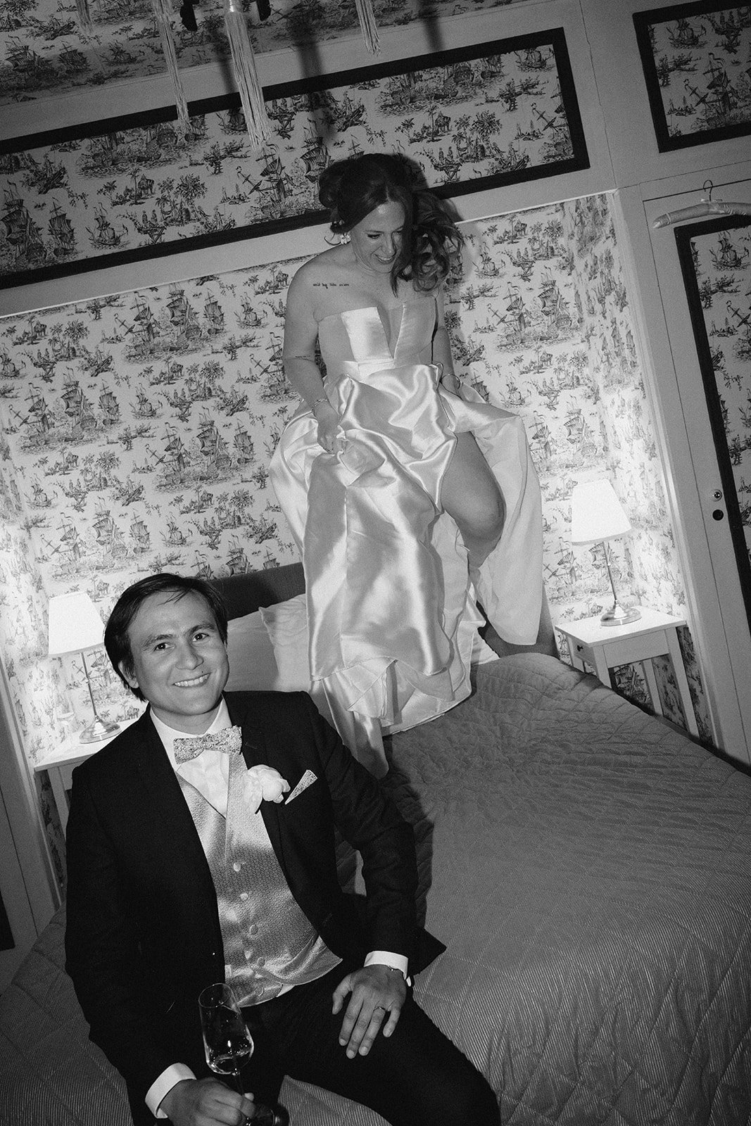 Une femme en robe de mariée se tient sur un lit, tandis qu'un homme en costume avec nœud papillon et boutonnière pose en face, tous deux souriants dans une chambre décorée.