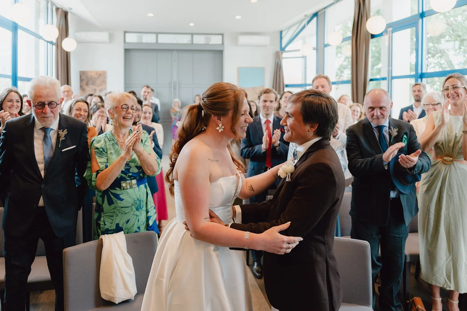 Un couple en mariage échangeant un regard à l'intérieur d'une salle, entourés de leurs invités qui applaudissent et sourient.