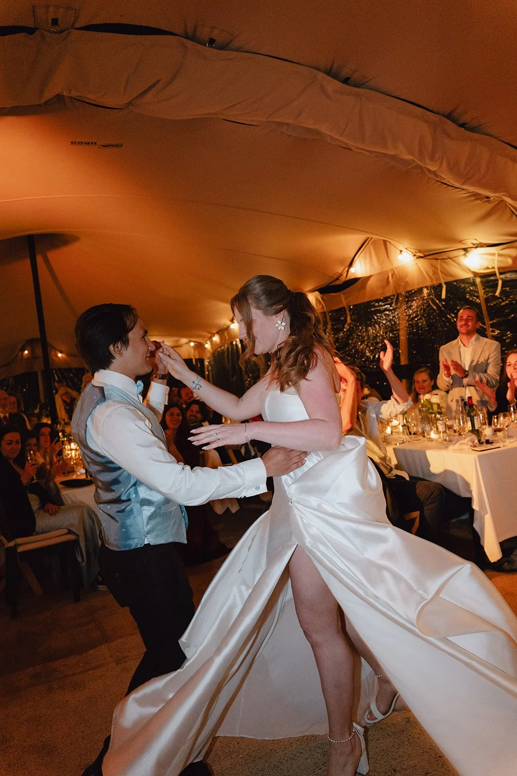 Un couple en robe de mariée et costume danse lors d'une soirée, entourés d'invités applaudissant. Caméra capturant un moment festif dans une tente décorée.