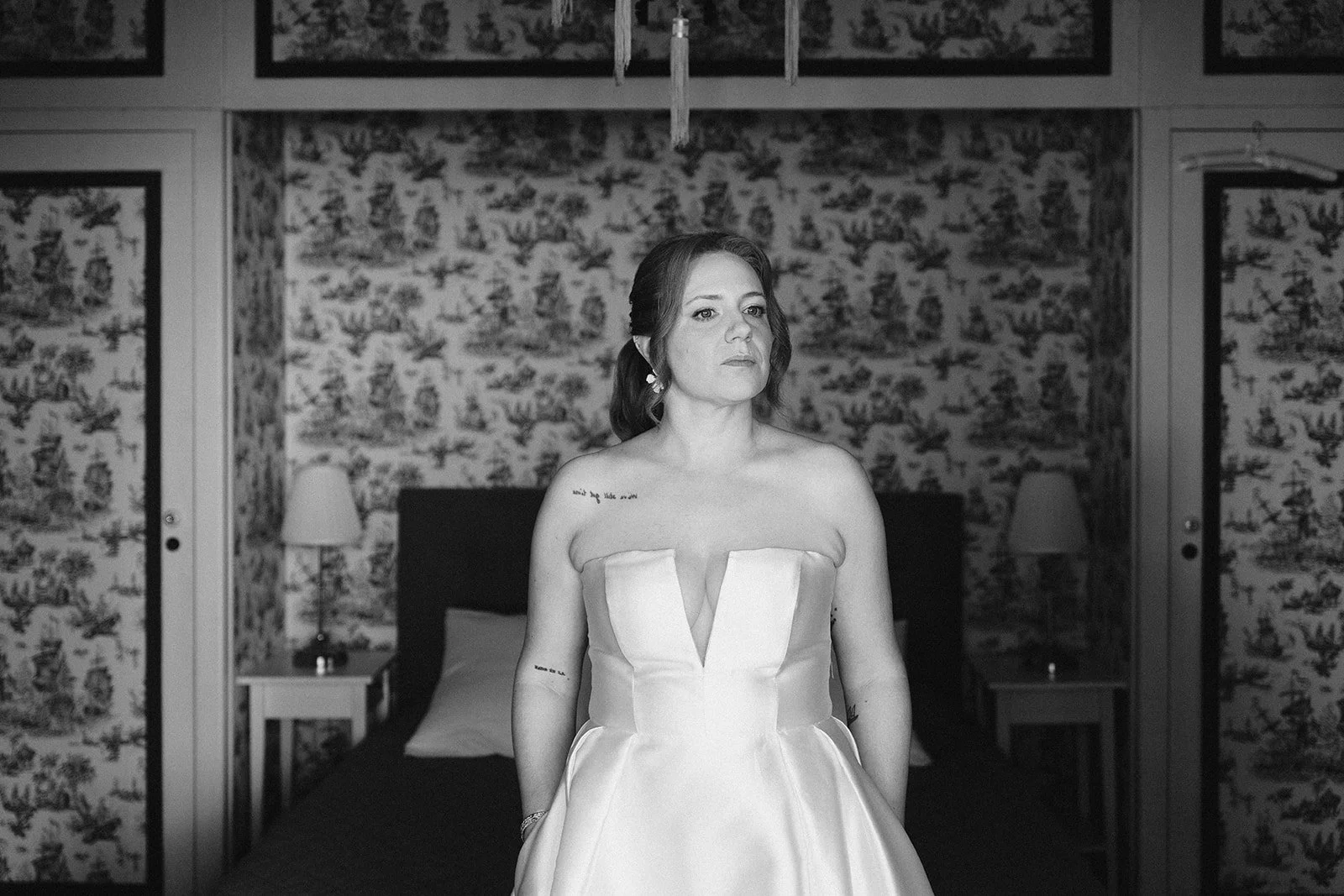 Une femme en robe de mariage assise sur un lit dans une chambre avec un décor floral, en noir et blanc, séance portrait mariée.