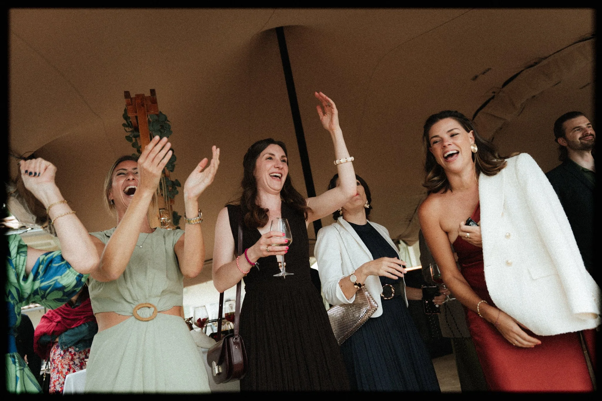 Groupe de personnes élargie, souriantes, lors d'une réception ou fête, tenant des verres de vin, sous un plafond en arc. Atmosphère festive et conviviale.