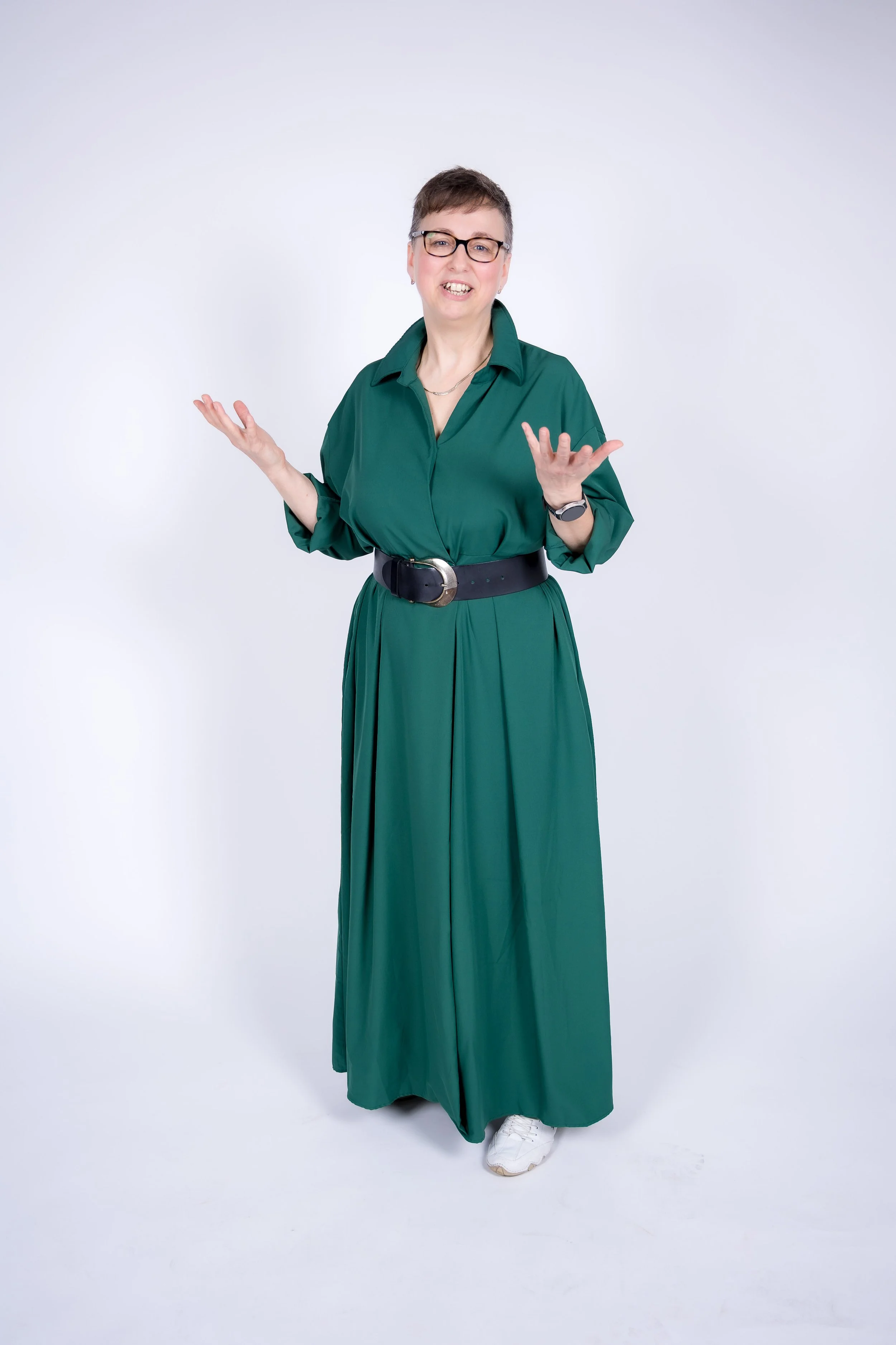 Eine Frau mit kurzen Haaren, Brille und einem grünen Kleid, die auf weißen Sportschuhen steht,  mit offener Geste und lächelt.
