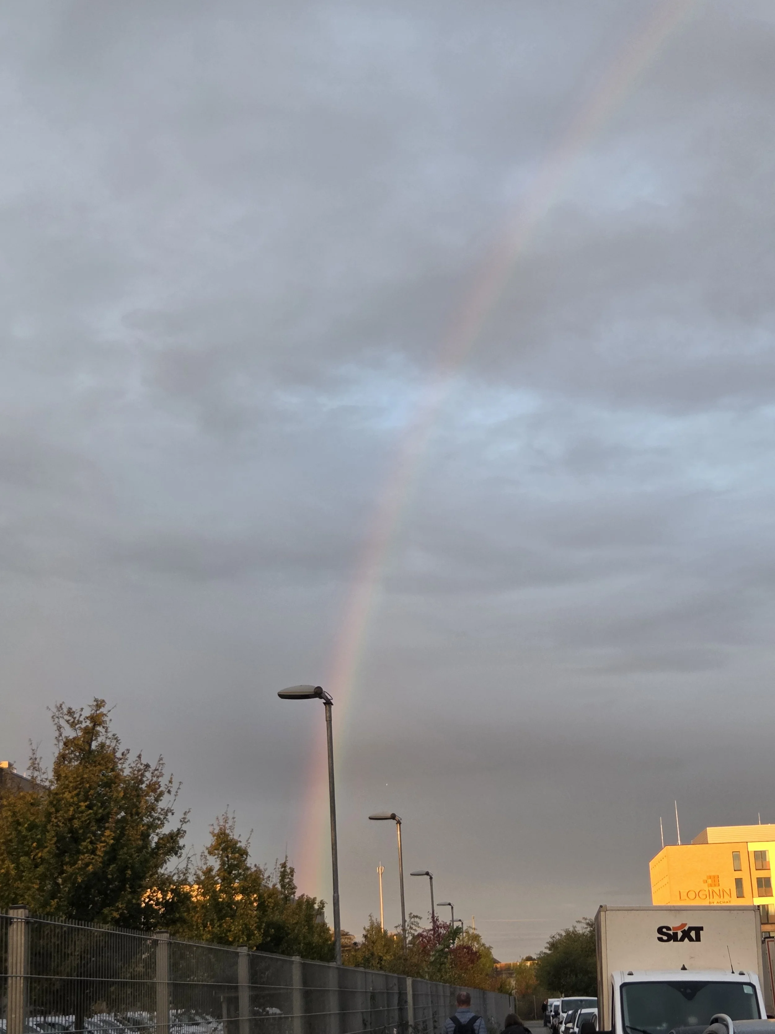 Regenbogen am Himmel über einem städtischen Parkplatz mit Bäumen, Straßenlaternen und Gebäuden, darunter ein gelbes Gebäude mit LOG-IN!