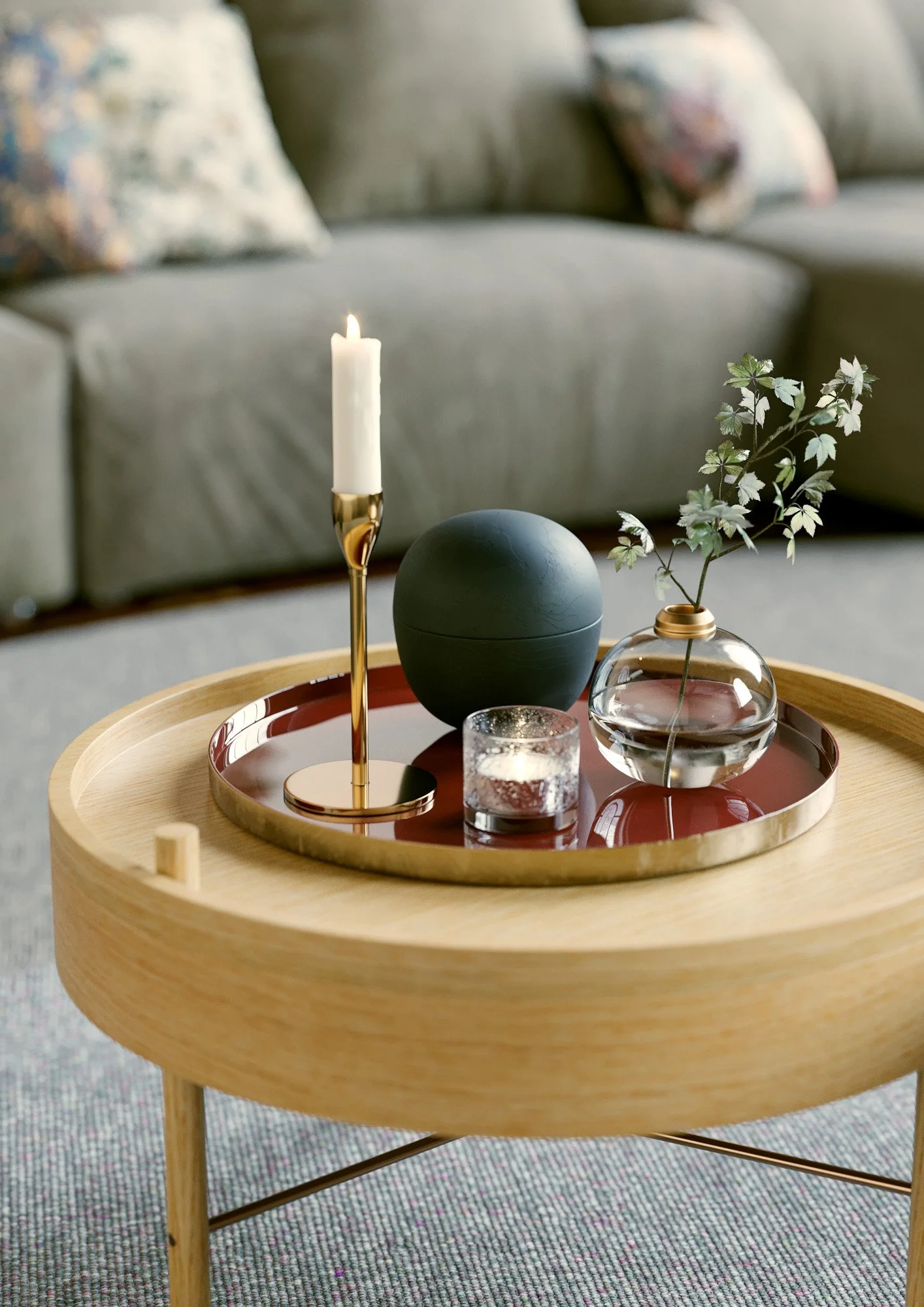 A wooden round side table with a tray holding a white lit candle in a gold candle holder, a green spherical object, a small glass candle holder with a candle, and a clear glass vase with green leafy branches. A gray sofa with patterned pillows is in