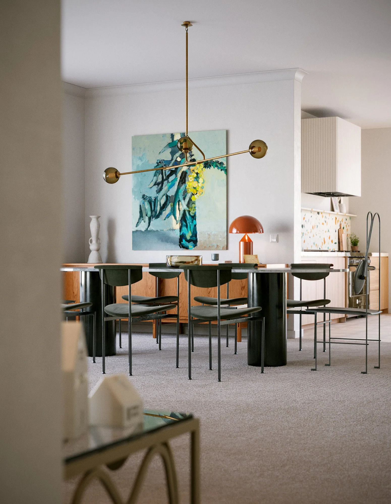 Modern dining area with a black round table, six black chairs, a colorful abstract painting on the white wall, a red and yellow table lamp, and a brass chandelier hanging from the ceiling.