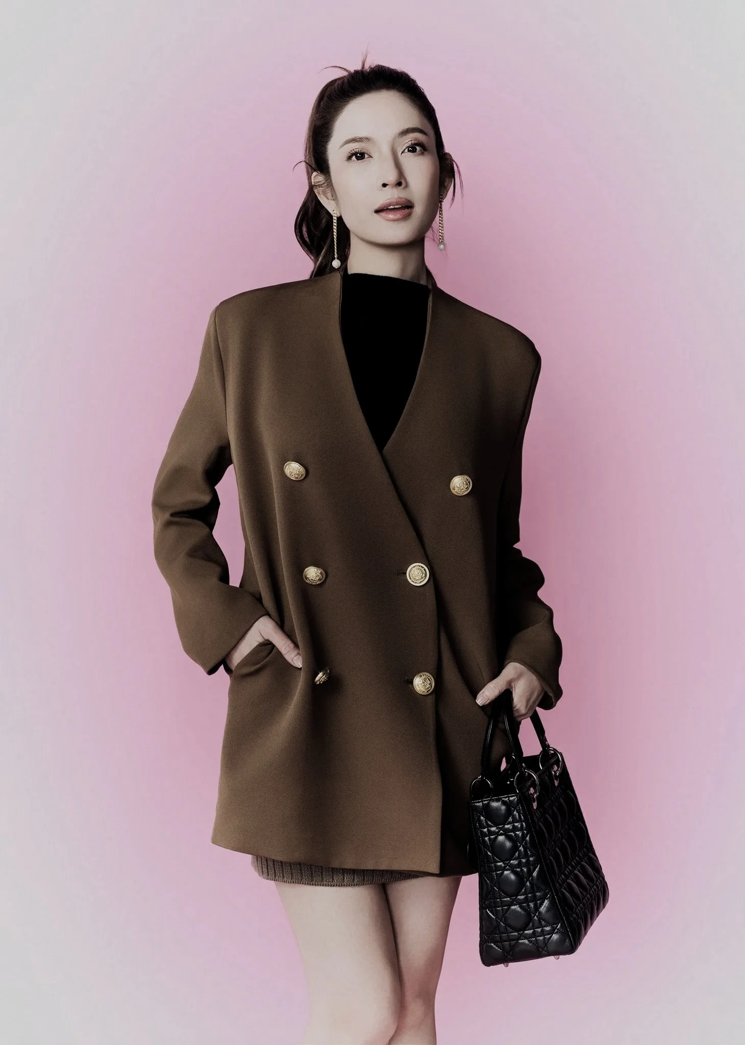 Woman wearing a brown double-breasted blazer with gold buttons, black top underneath, holding a black quilted handbag, standing against a pink background.