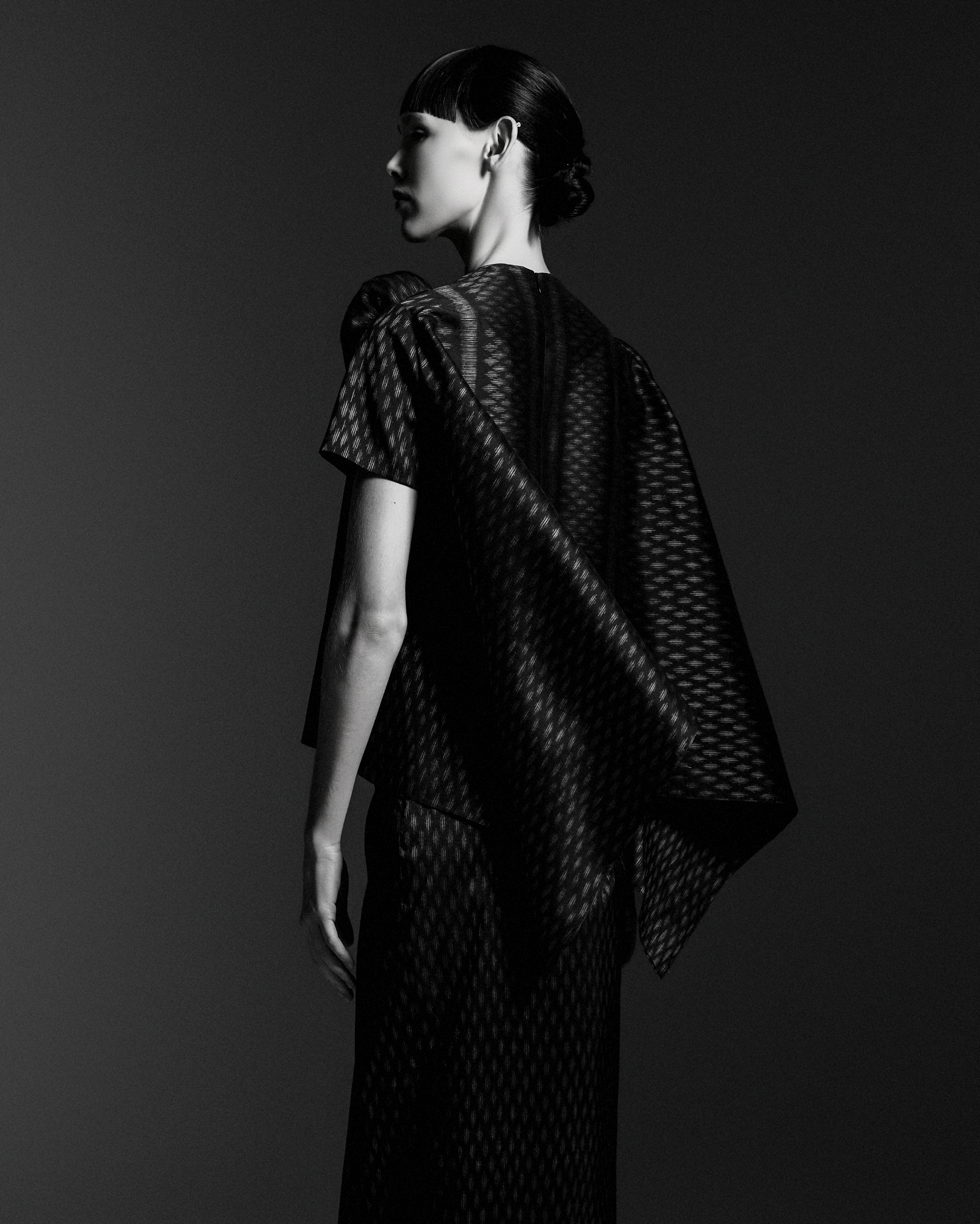 A black-and-white photograph of a woman with short dark hair styled in a sleek, shiny bob with bangs. She is looking to her left, and her face is in profile. She is wearing a sheer, patterned dress with short sleeves, and the background is plain and 