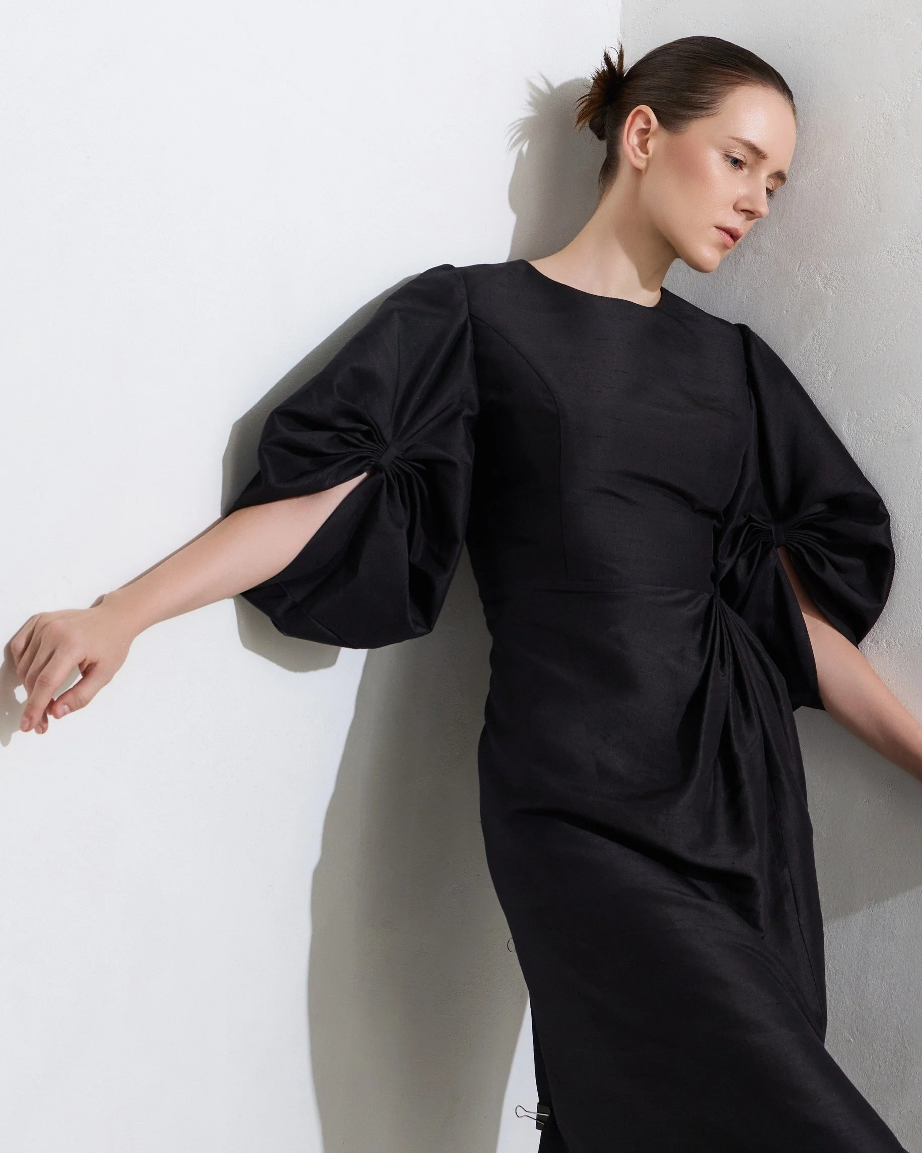 A woman with brown hair in a ponytail wearing a black dress with puffed sleeves, leaning against a white textured wall.