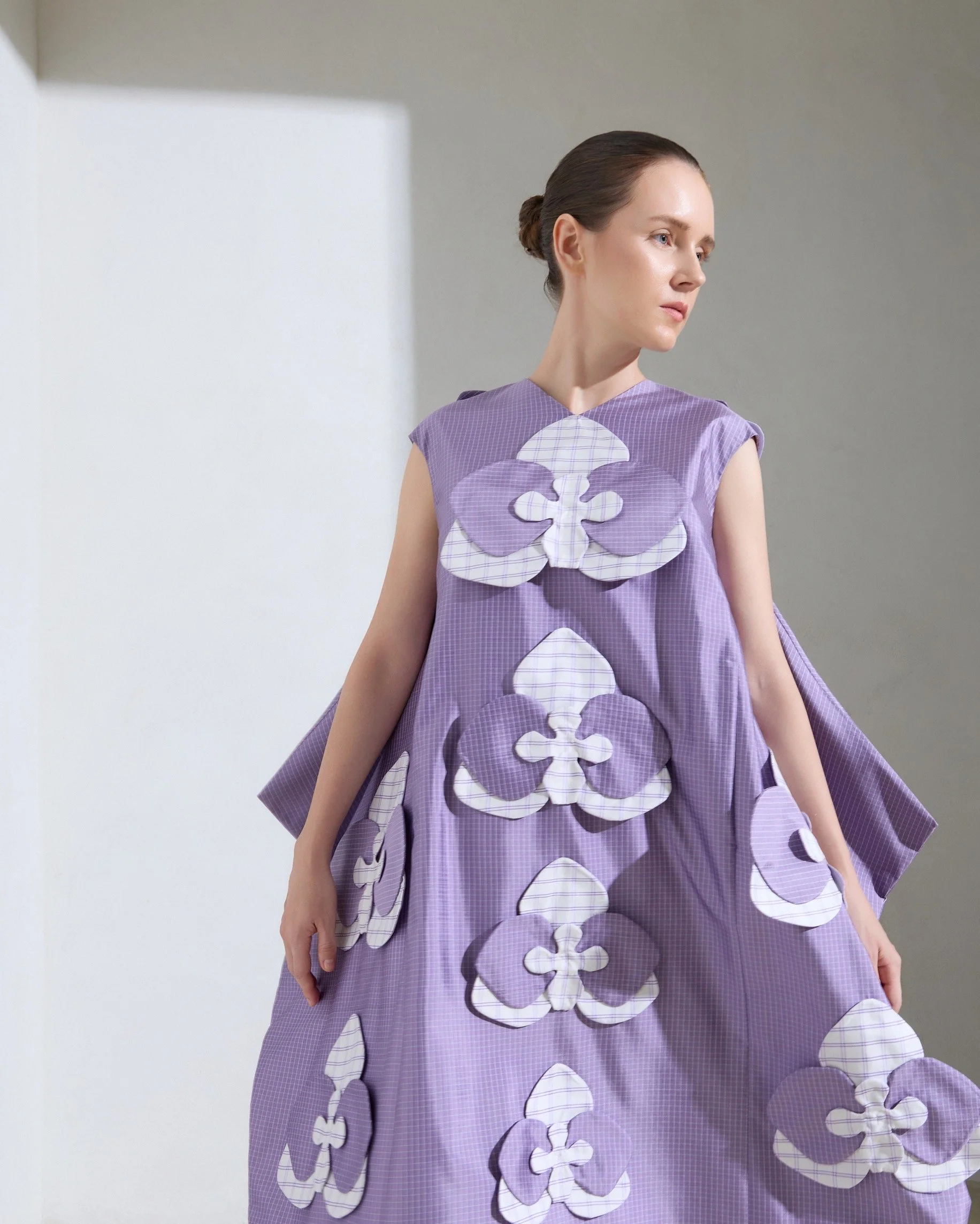 Woman in a purple dress with butterfly-shaped patterns and white checked details, standing against a plain wall.