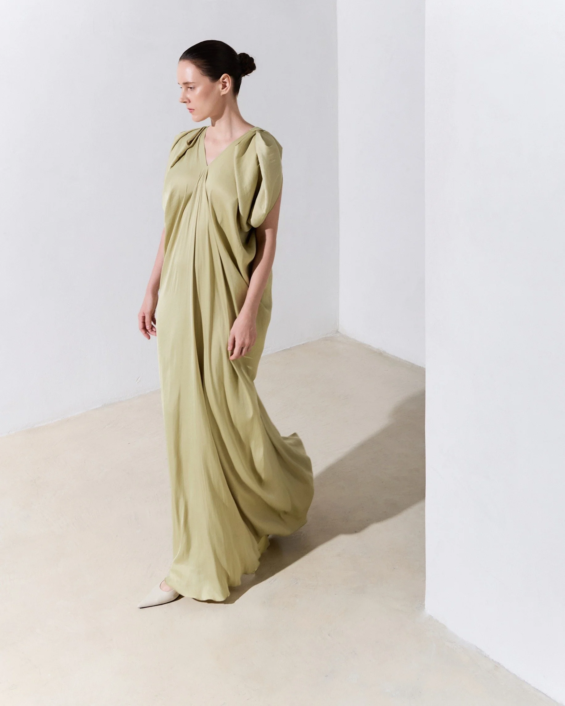 A woman with dark hair tied back, wearing a flowing, light yellow dress with puffed sleeves, standing in a minimalist white room with a corner, looking downward and slightly to the side.