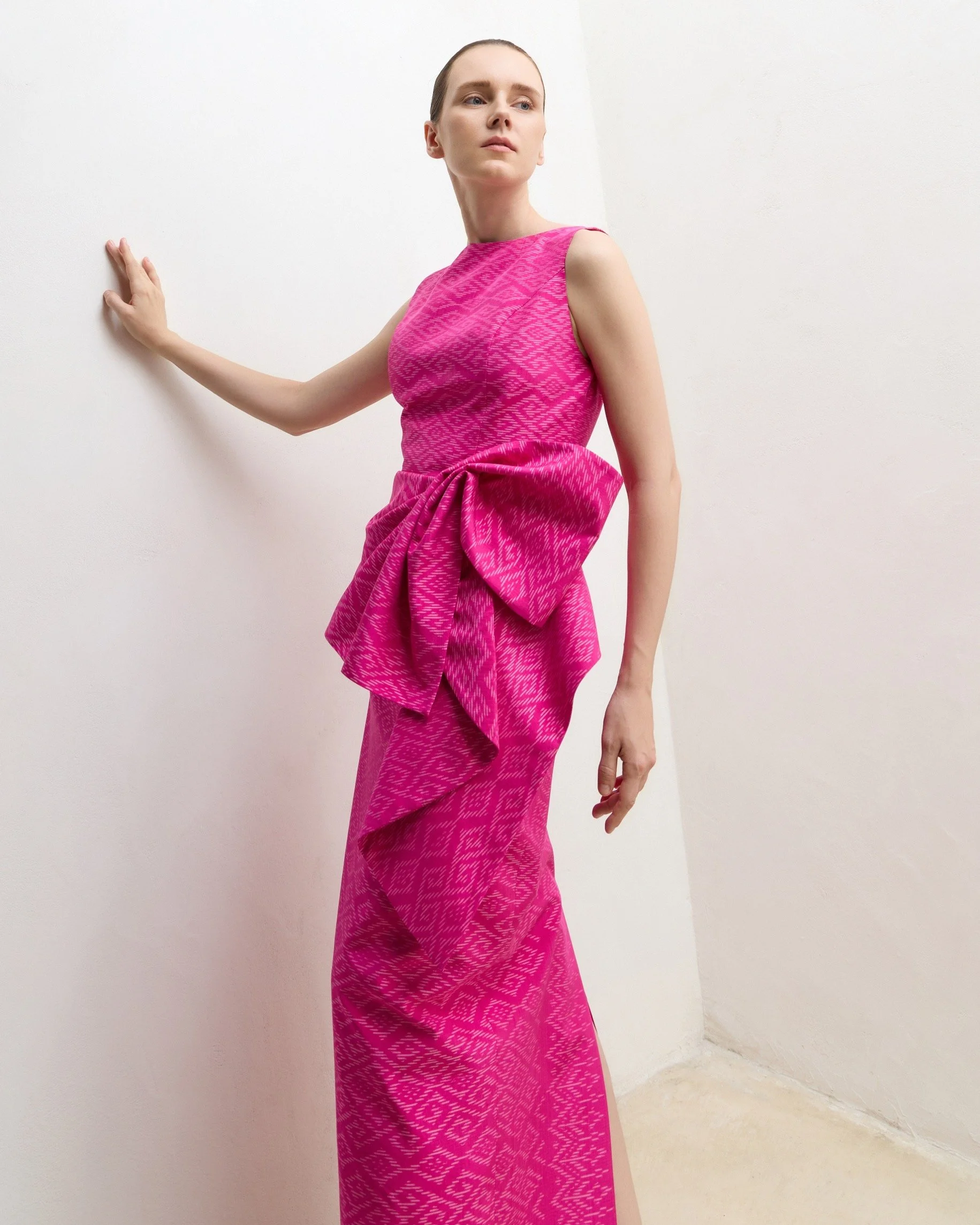 A woman wearing a pink patterned sleeveless dress with a large bow around the waist, standing against a white wall.
