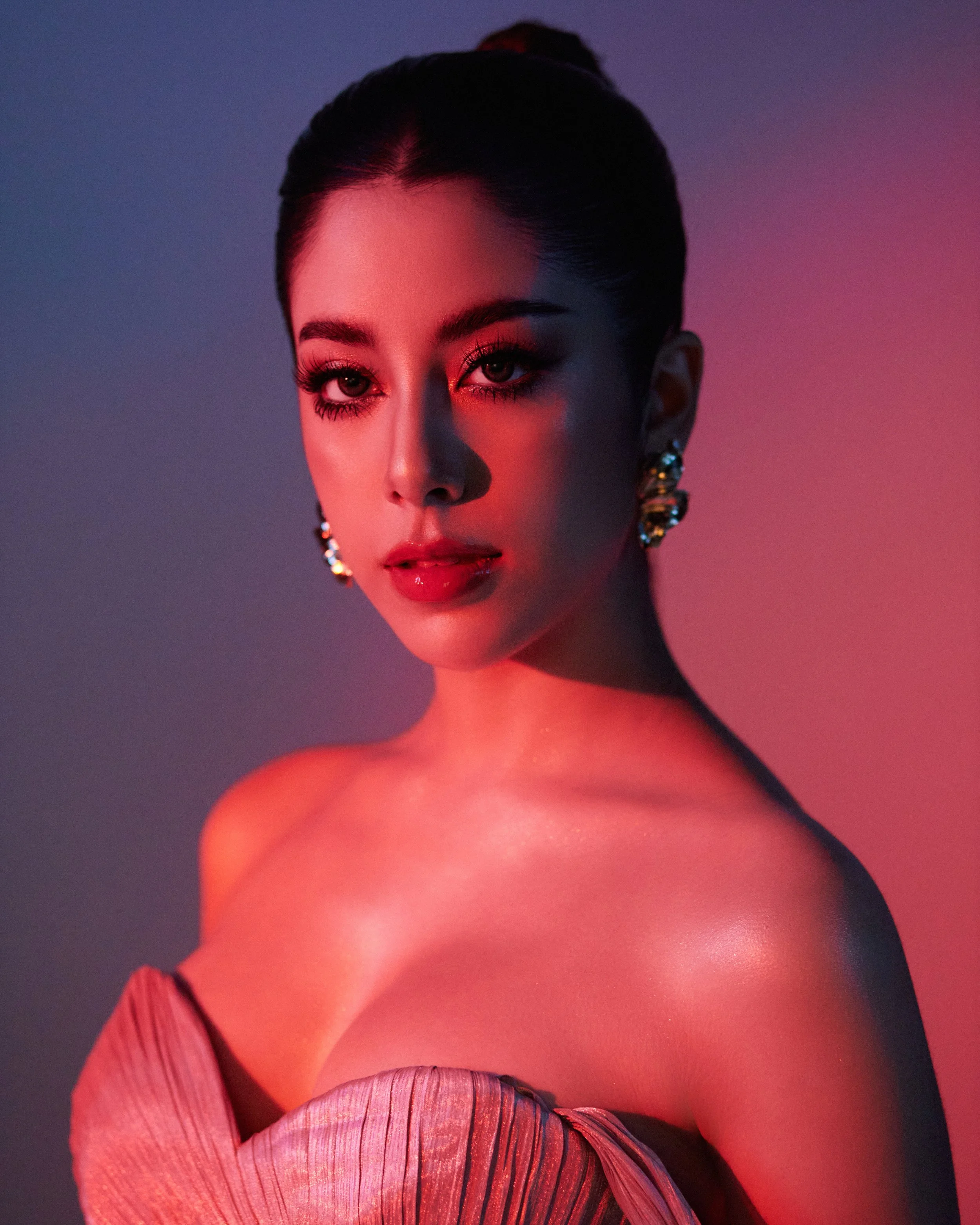 Close-up of a woman with dark hair in an elegant updo, wearing bold makeup with dark eye makeup and red lipstick, adorned with earrings, with a background of soft purple and pink lighting.