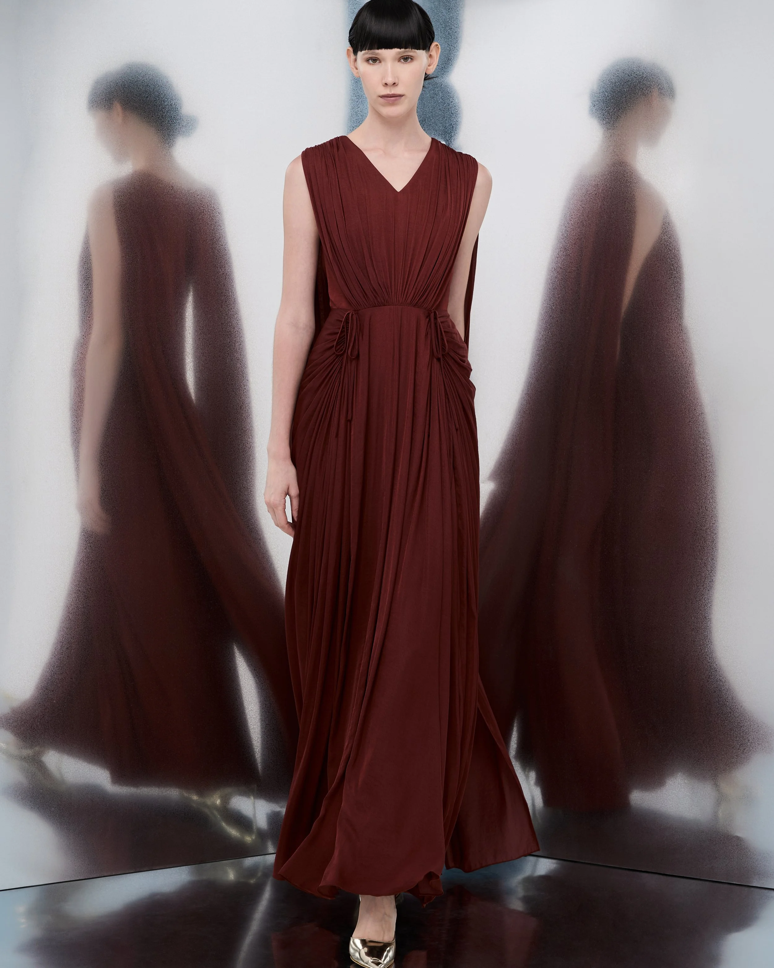 A woman in a long red pleated dress with shoulder cutouts and a waist tie stands against a reflective metallic wall. Her reflection appears blurred and distorted on the wall.