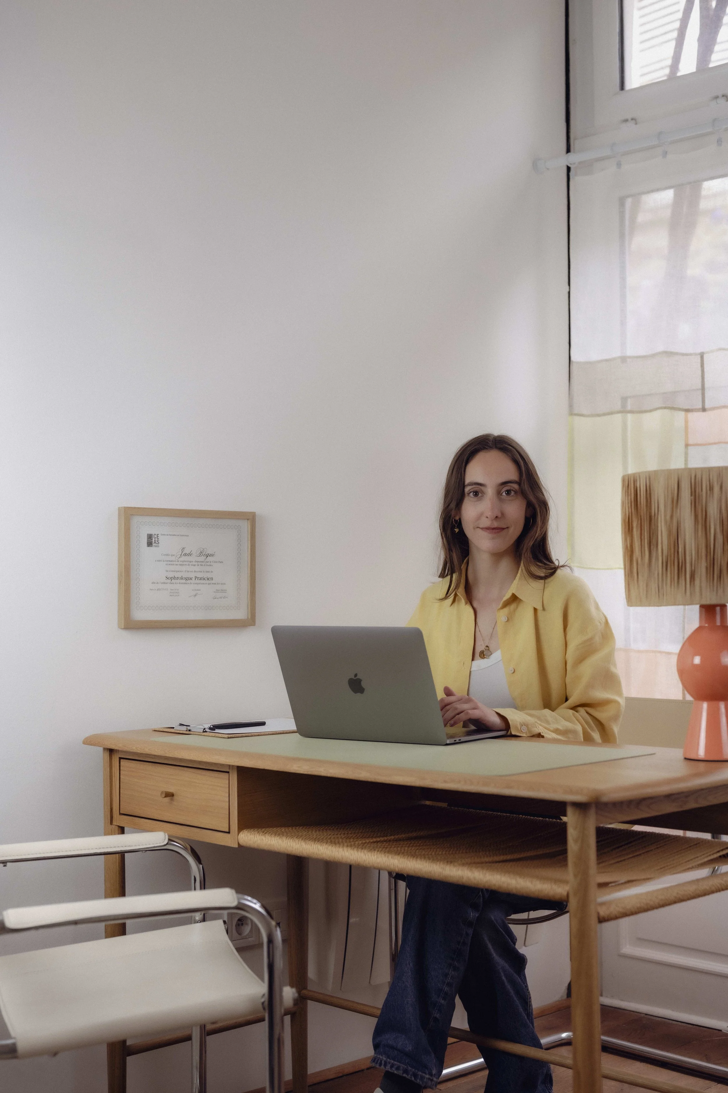 Femme assise à un bureau en bois, utilisant un ordinateur portable, dans une pièce lumineuse avec une lampe orange et une fenêtre avec rideaux colorés.