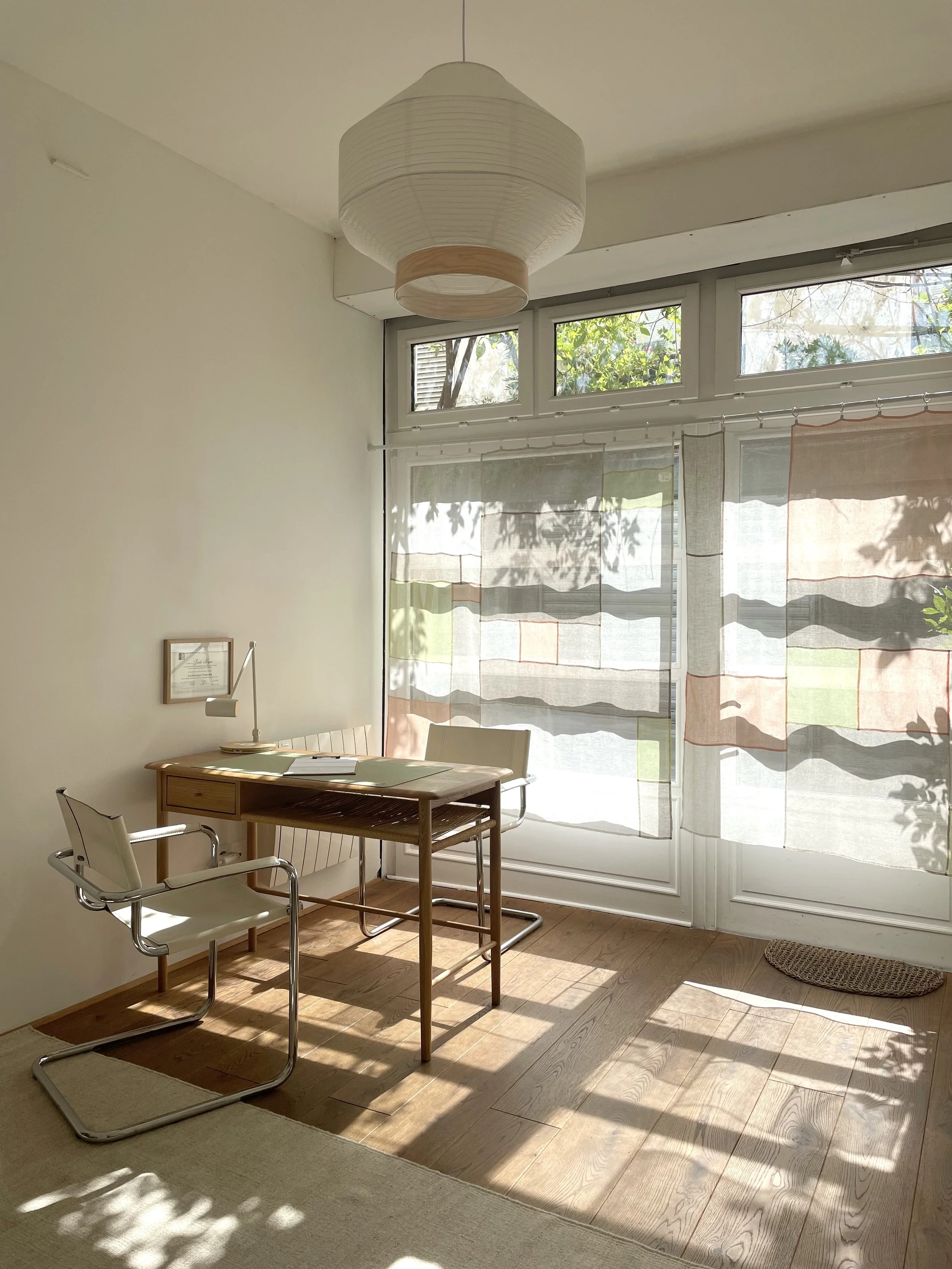 Une pièce lumineuse avec une table en bois, deux chaises en métal et en cuir, une grande fenêtre avec des rideaux semi-transparents. Un plafond avec un luminaire en papier rond.